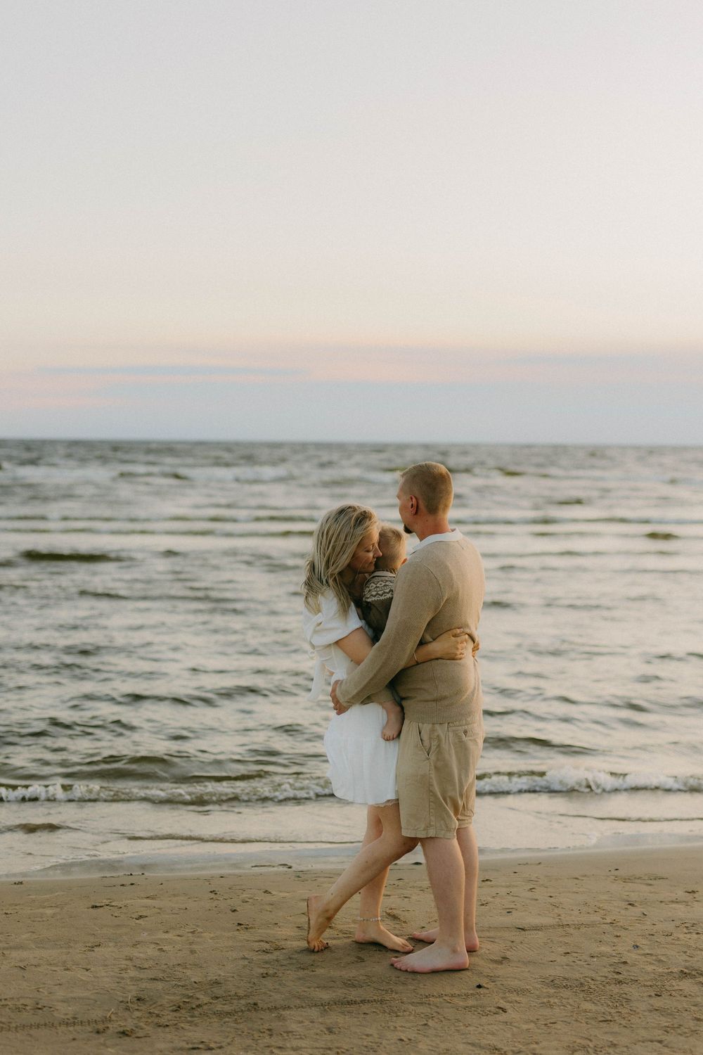 A couple embraces while standing on a beach at sunset with waves rolling in the background.