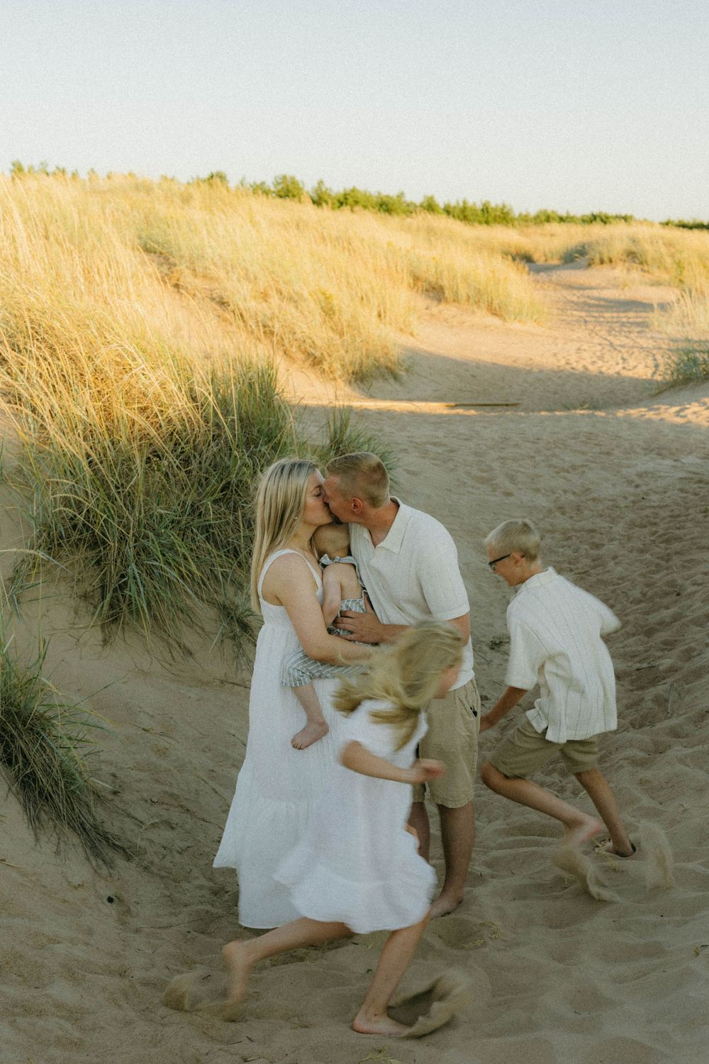 A group in light summer clothing plays together on sandy beach dunes during a warm golden hour.