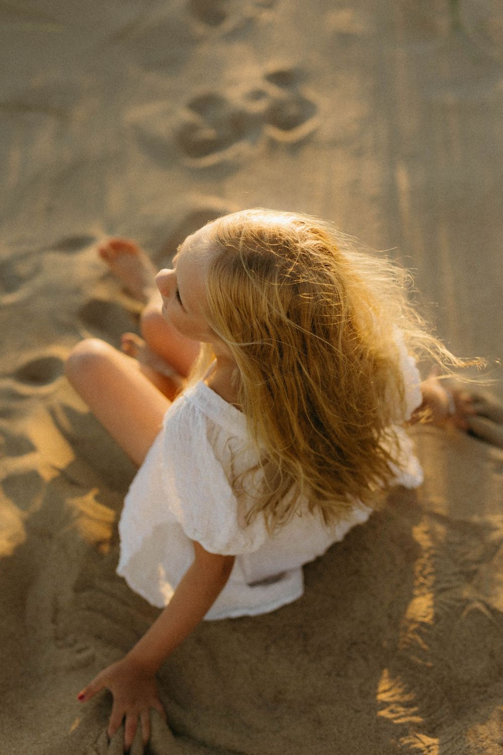 A young child in a white dress plays in the sand at golden hour, with sunlit blonde hair flowing in the breeze.