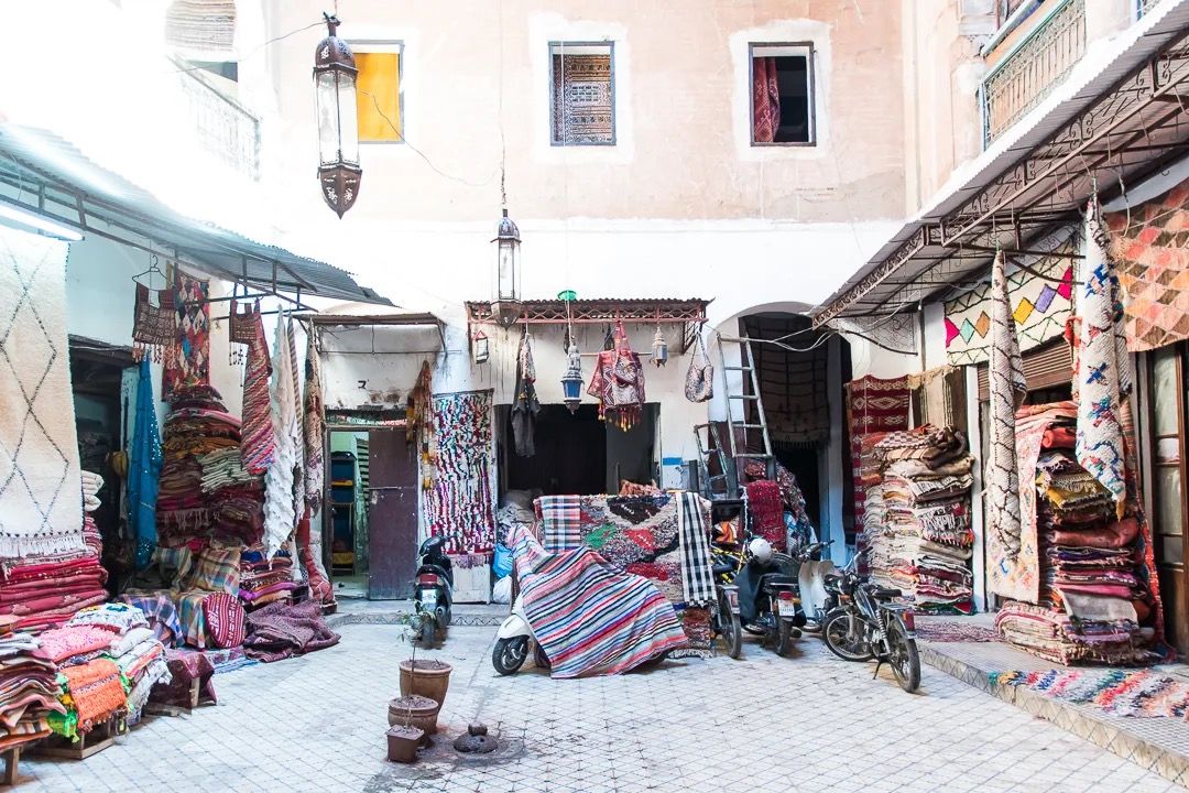 A colourful Moroccan market street lined with hanging textiles, lamps and wooden stalls.
