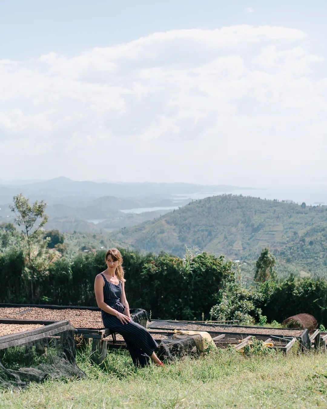 Woman sitting on a wooden bench surrounded by greenery with rolling mountains in the distance.