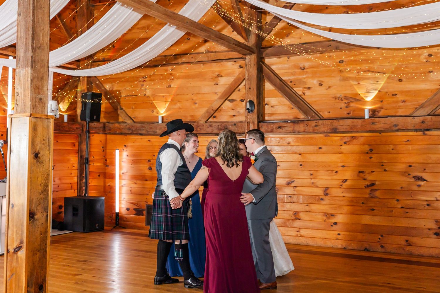 Couples dancing together in a rustic wooden barn venue decorated with white fabric draping across the ceiling.
