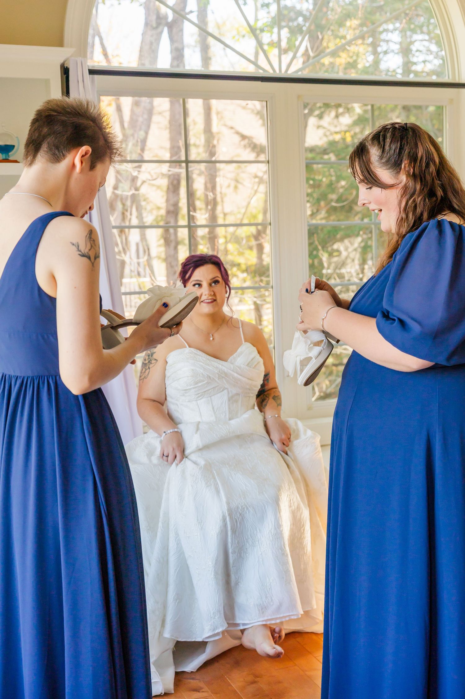 Bridesmaids in royal blue dresses help adjust a white wedding gown in a sunlit room.