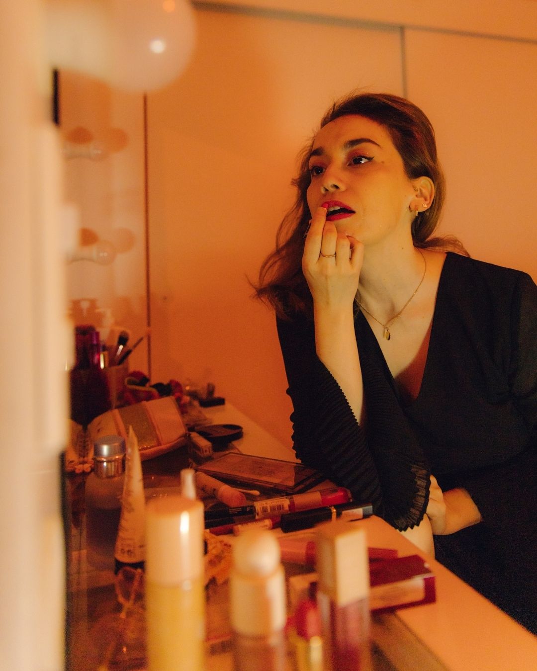 A woman applies red lipstick while looking in a mirror surrounded by candles and makeup products.