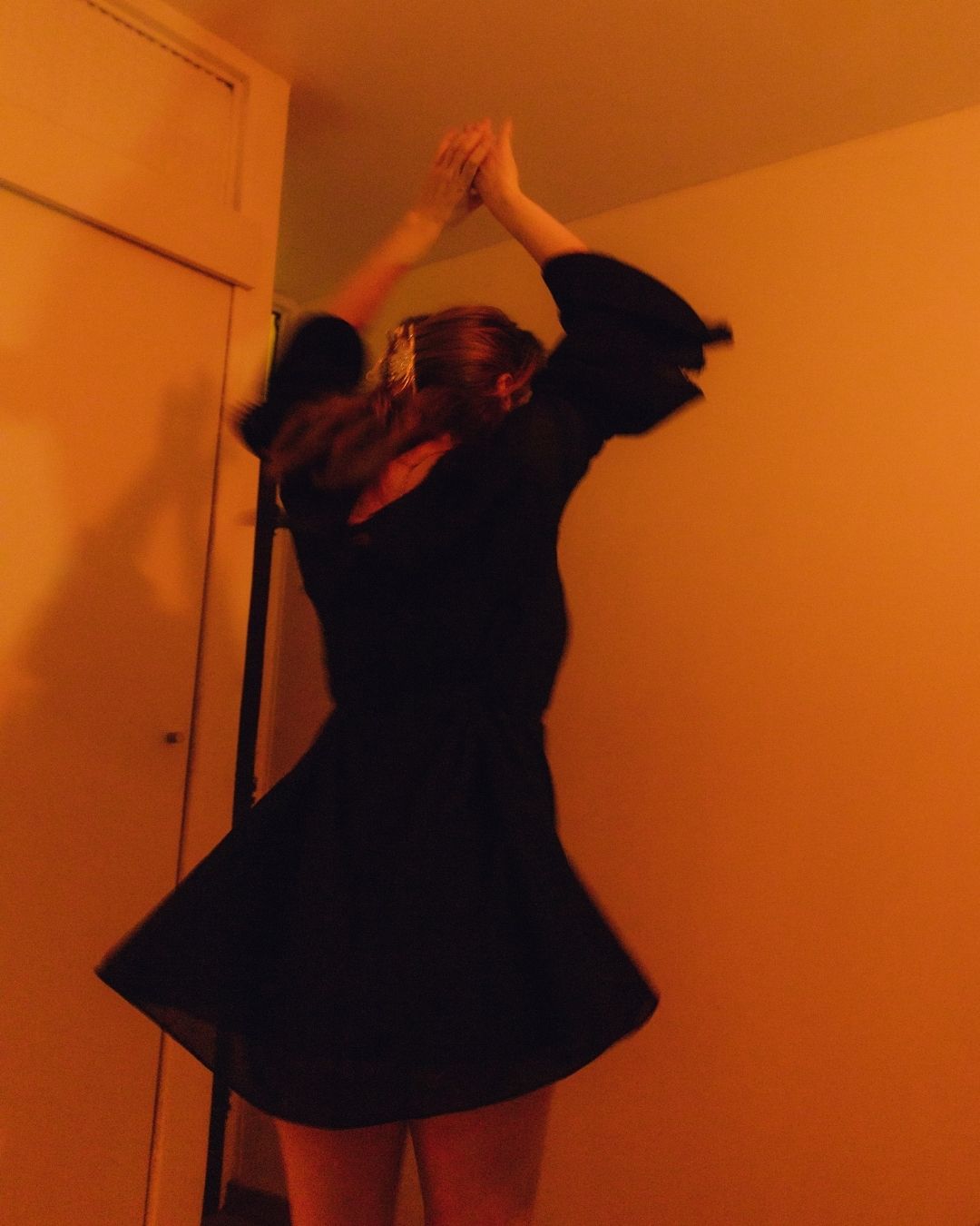 A woman in a black flared dress stretches her arms upward against an orange-toned wall.