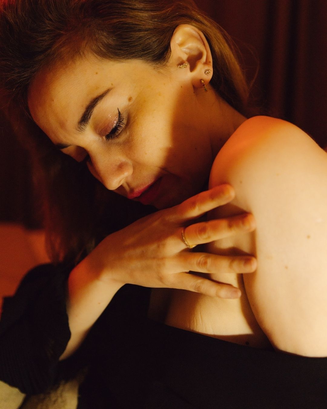 A woman in a black top gently touches her bare shoulder while looking down in soft warm candlelight.