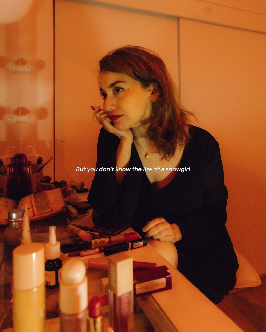 A woman sits thoughtfully at a vanity mirror with makeup products, with text overlay about a showgirl.