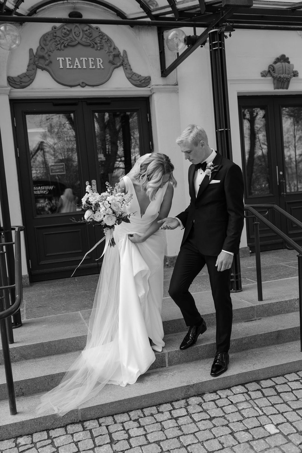 A wedding party walking down a sunny street in formal attire with bouquets of white flowers.