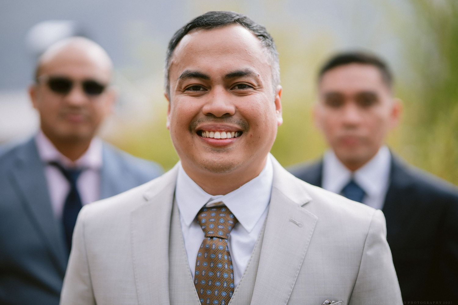 Groomsmen in light gray suits smile during an outdoor wedding.