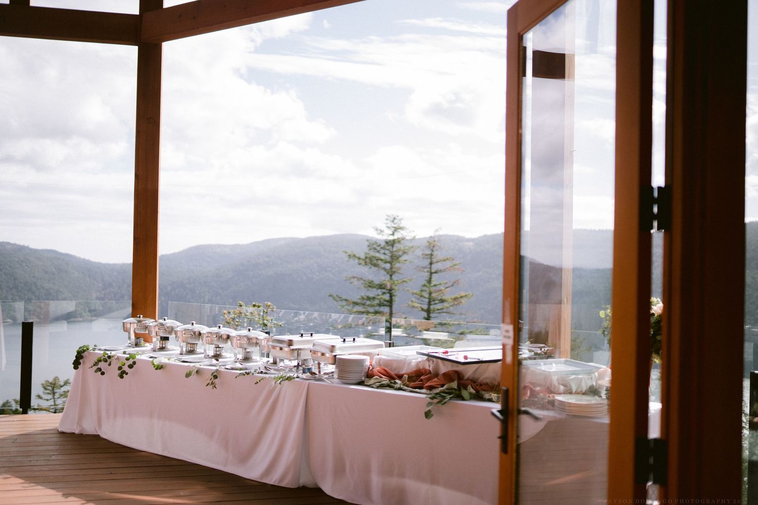 Long buffet table with white linens set up on outdoor deck overlooking mountain lake vista.