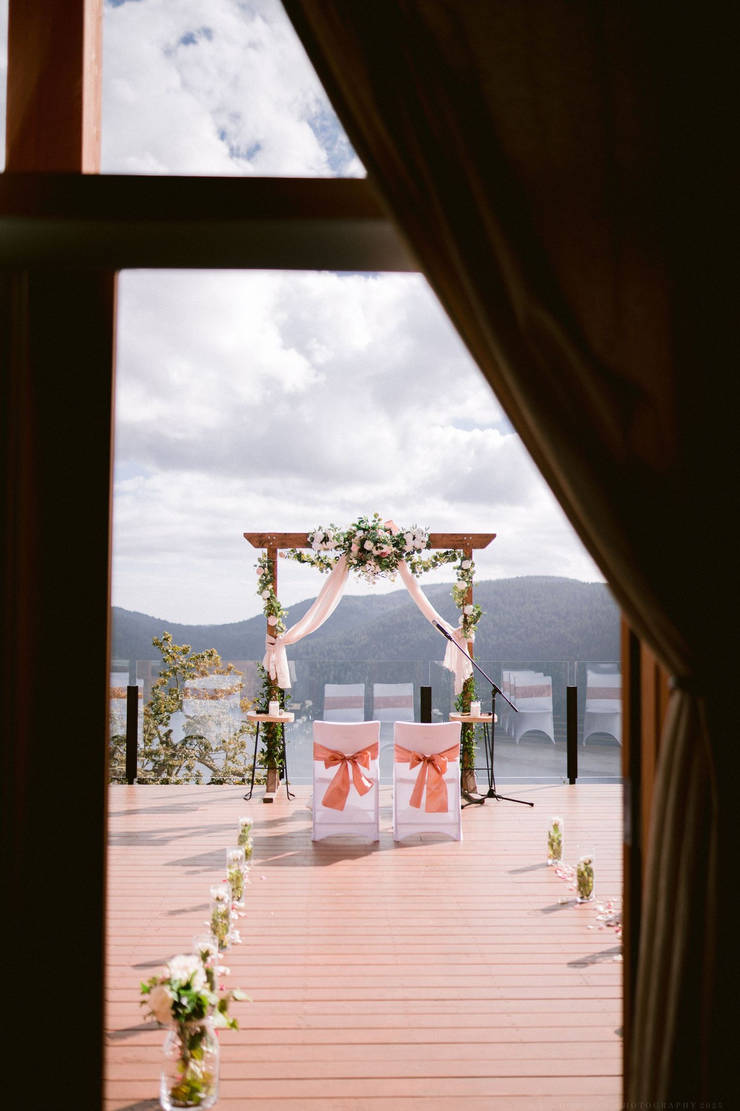 An outdoor wedding ceremony setup with floral arch and white chairs overlooking a mountain lake.