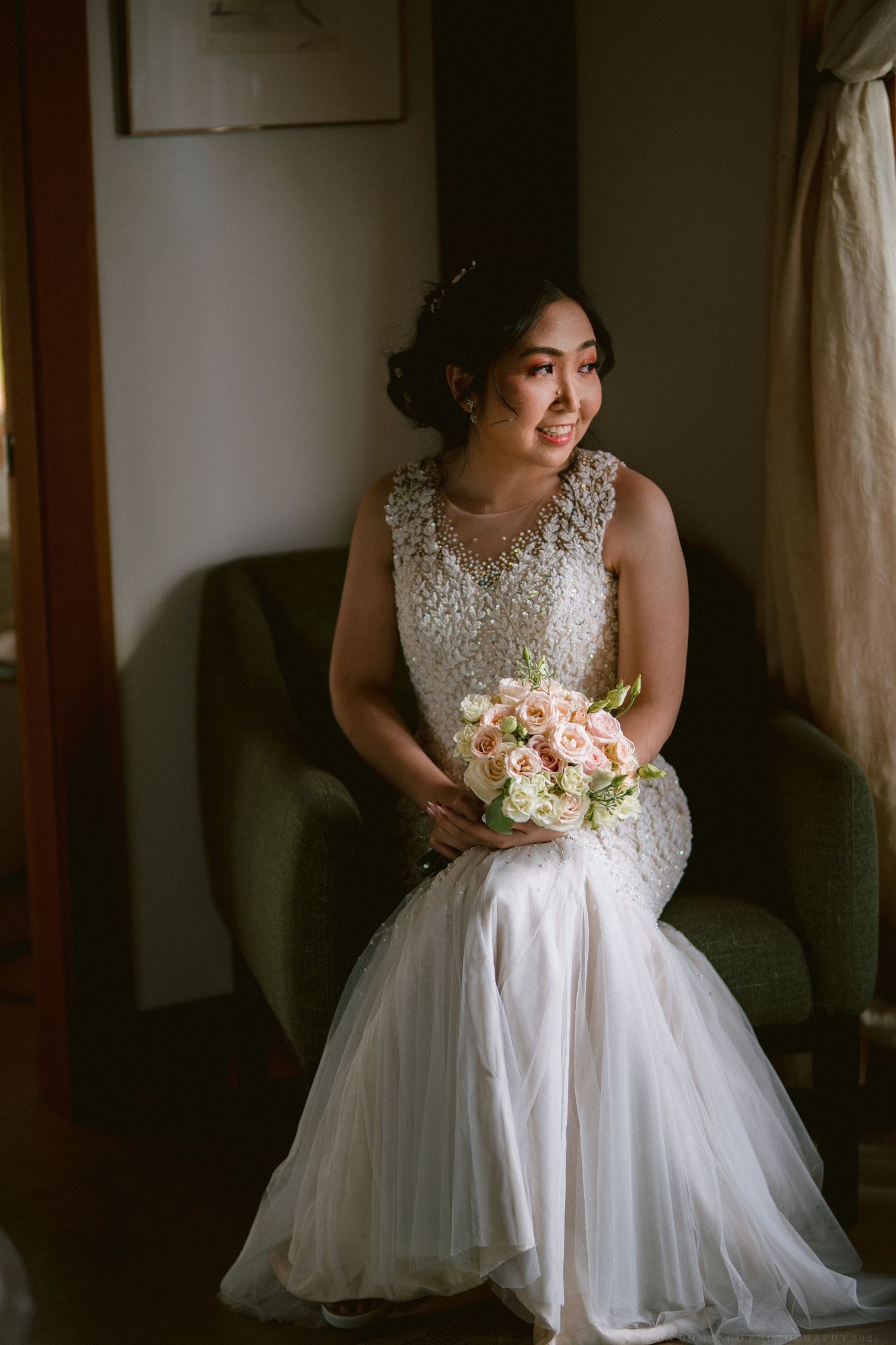 A bride in an elegant white gown sits holding a bouquet in an indoor setting with natural light.