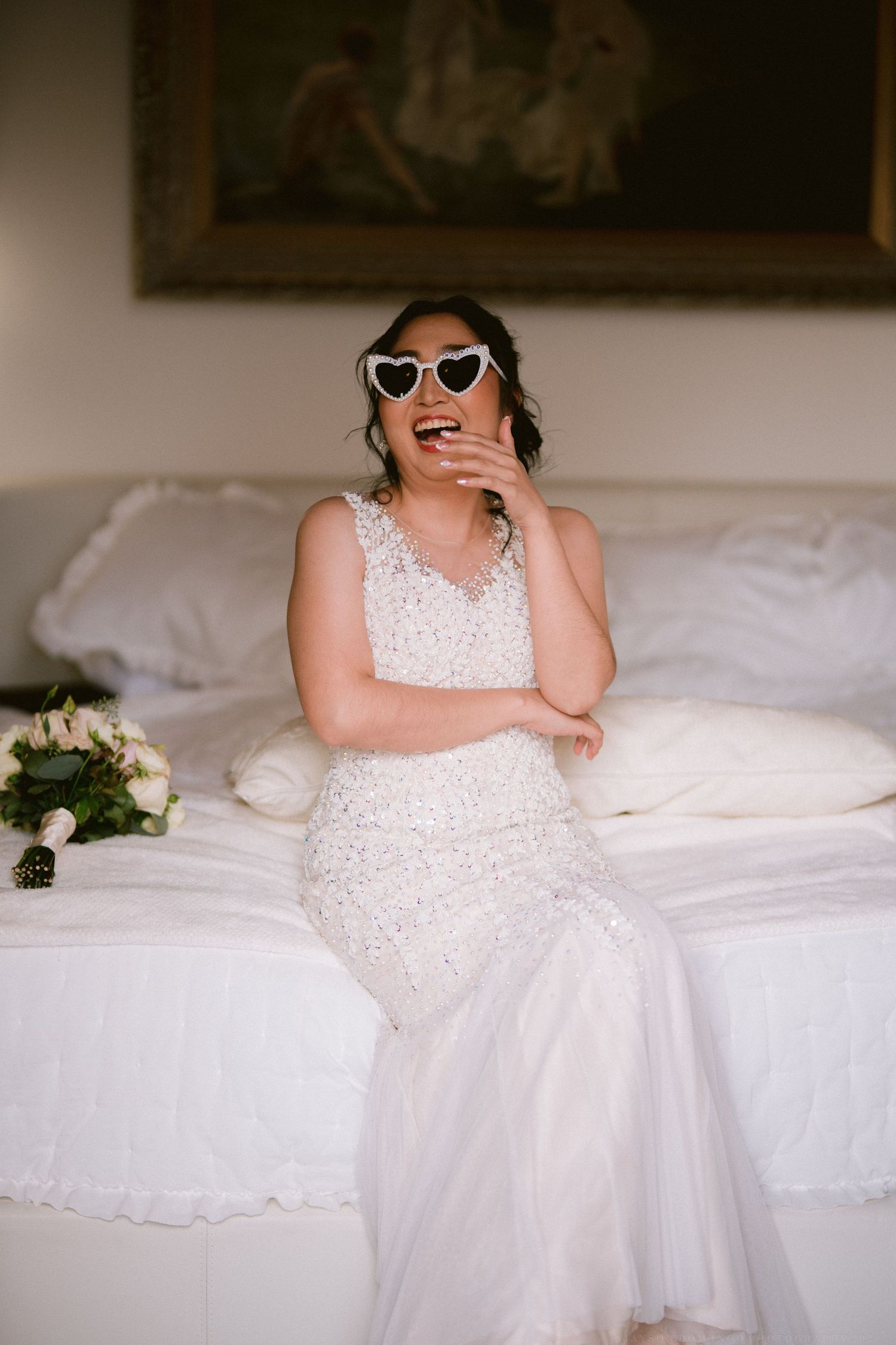 Playful bridal boudoir photos on white bedding featuring retro sunglasses and elegant white gown details.