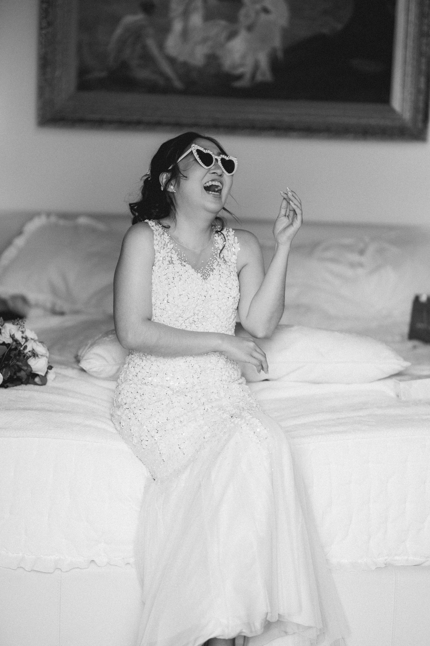 A bride in a white beaded dress reclines on white bedding while laughing.