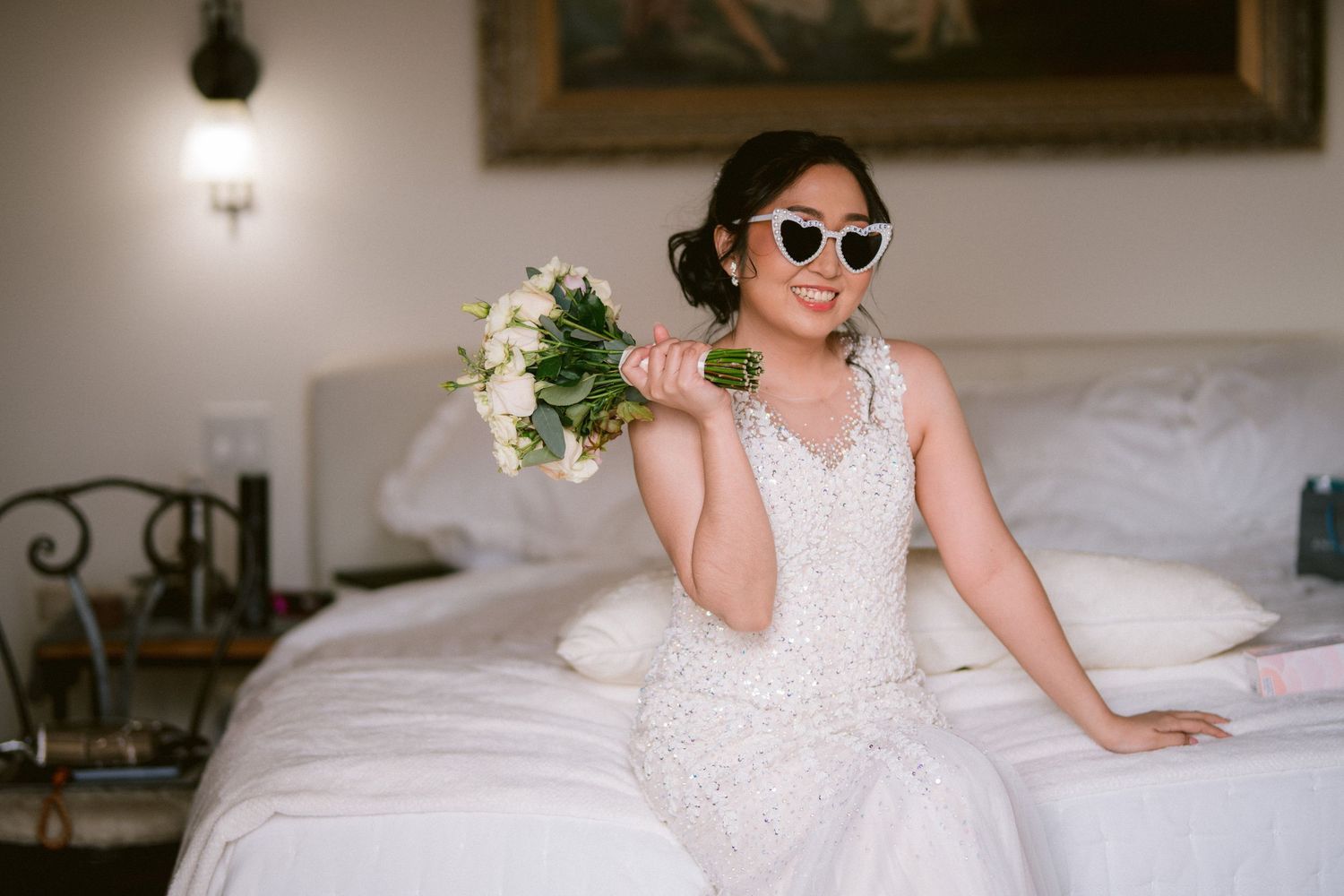 A bride relaxes on a white couch wearing sunglasses and a lace wedding gown.