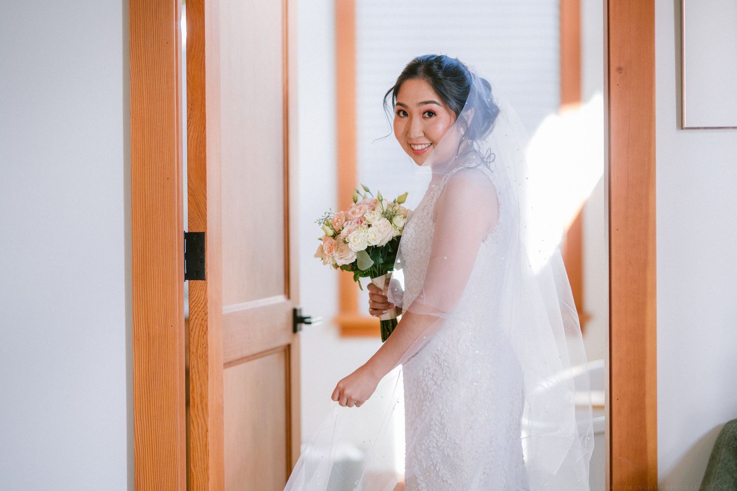 A person wearing a white wedding dress and veil holds a bouquet while standing in a doorway.