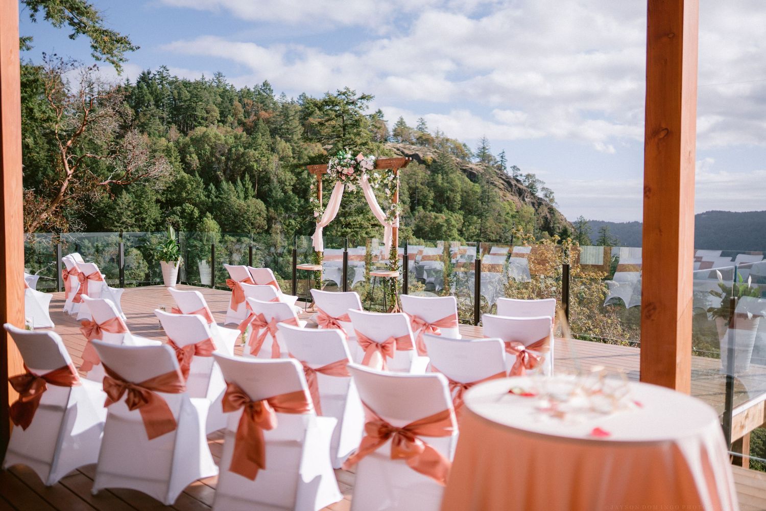 Outdoor wedding ceremony setup with white chairs and coral sashes overlooking mountain views.