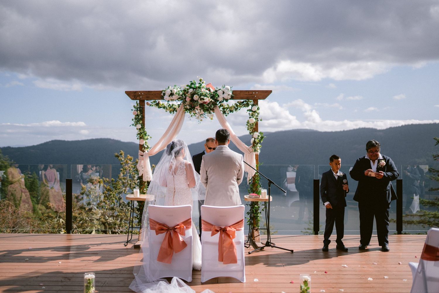Wedding ceremony under a floral arch with mountain views and cloudy skies in the background.