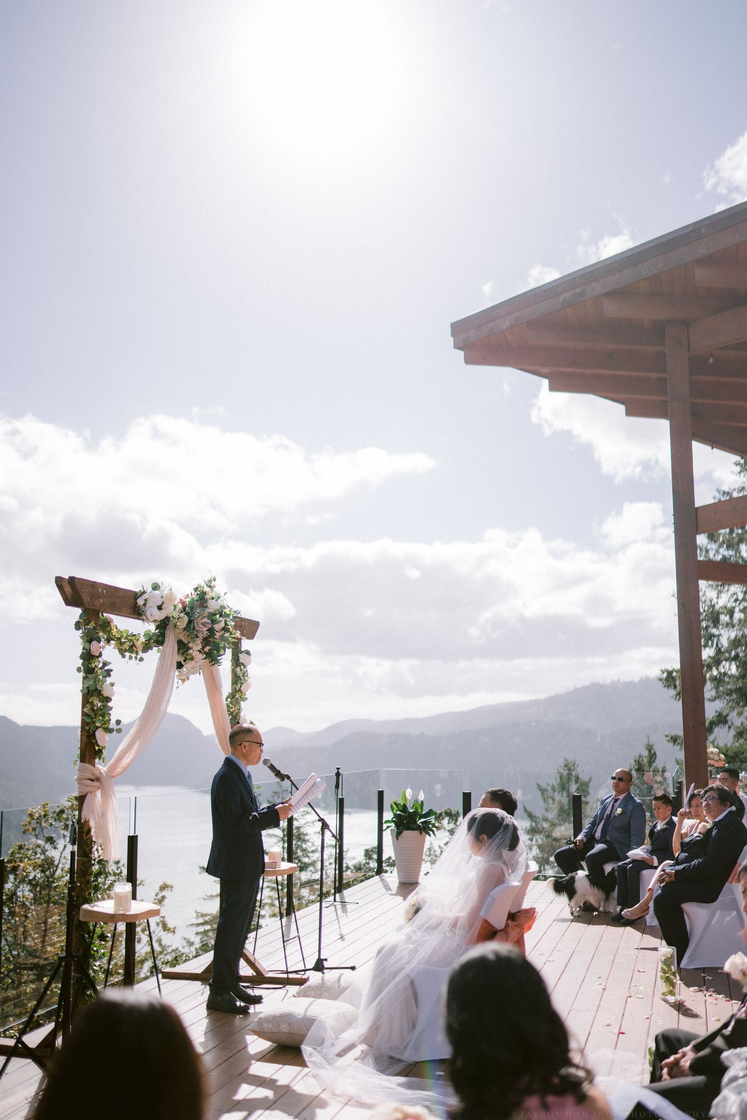 Scenic mountain wedding ceremony on deck under wooden arch with gathered guests.