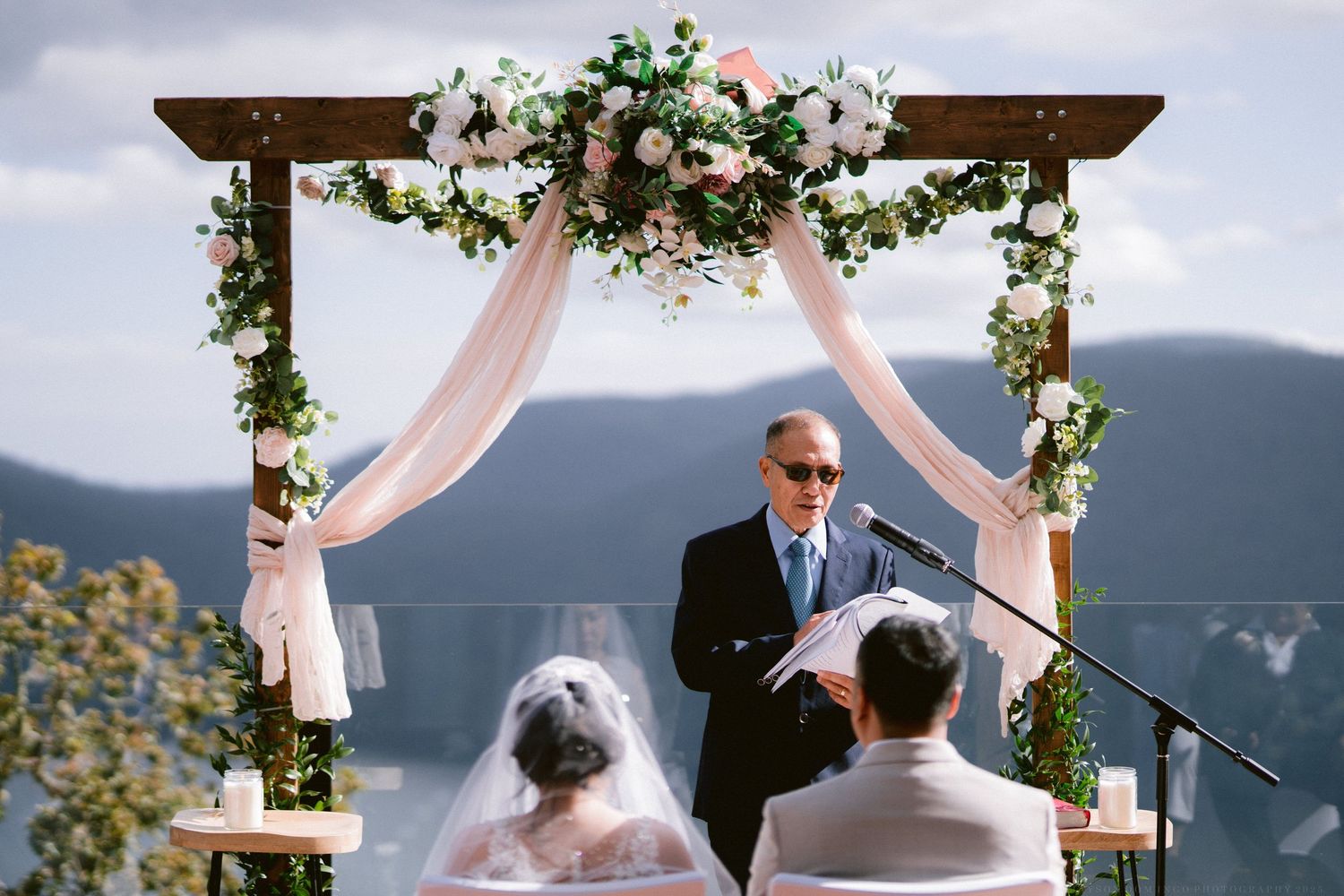 Outdoor wedding ceremony setup with floral arch and pink draping against a mountain backdrop.