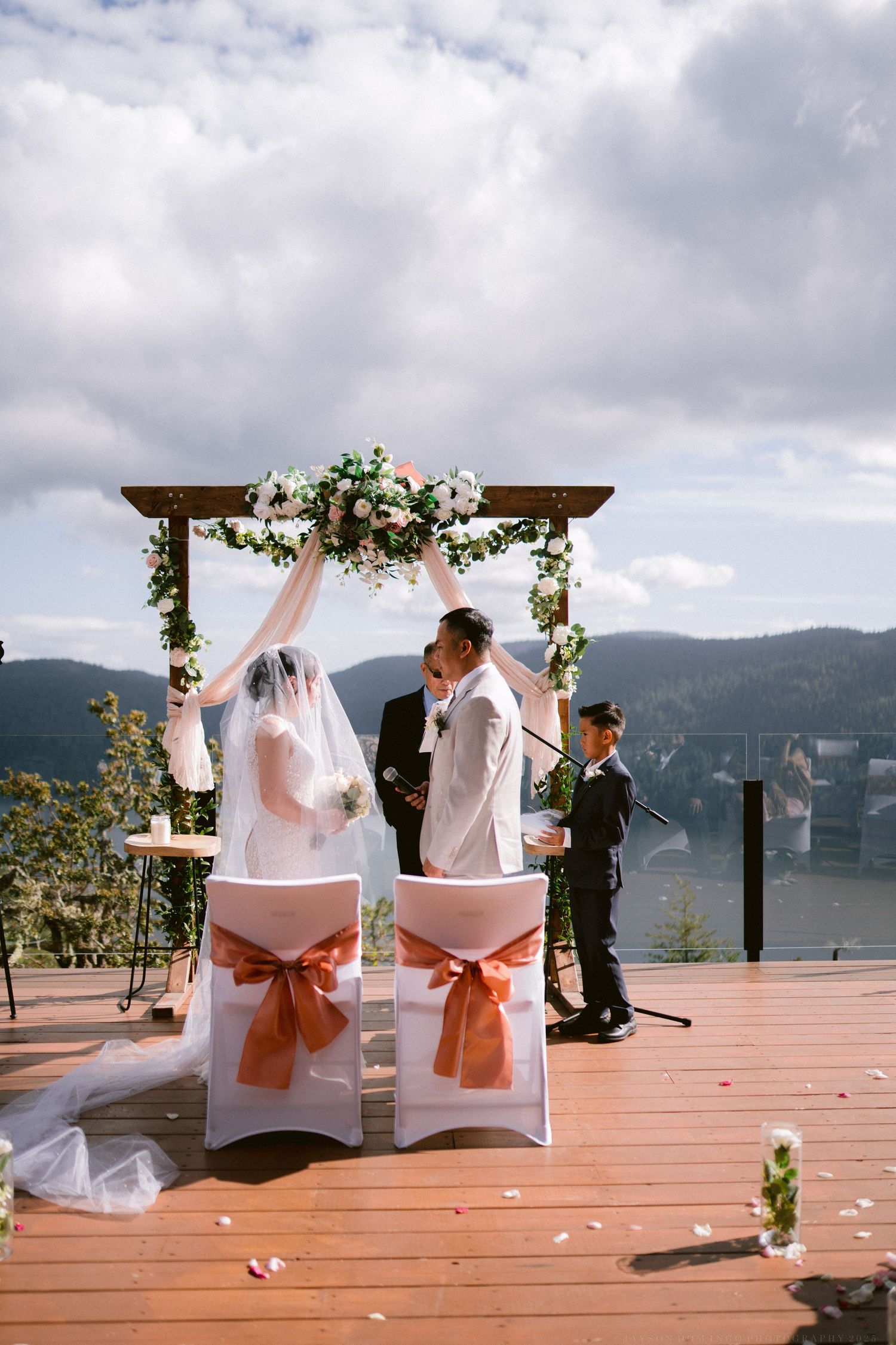 Outdoor wedding ceremony setup on a deck overlooking mountains with white chairs and floral arch decorations.