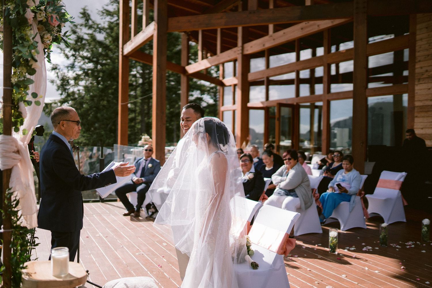 Outdoor wedding ceremony at a wooden lodge venue with guests seated on white chairs.