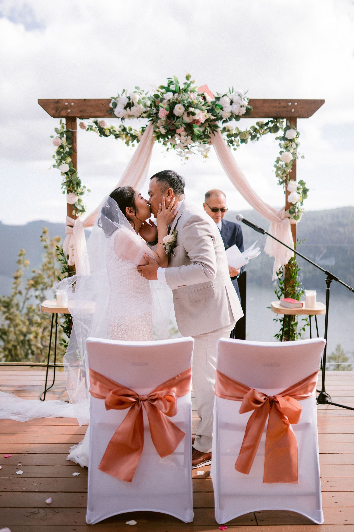 White ceremony chairs adorned with coral sashes arranged on a deck overlooking mountains.