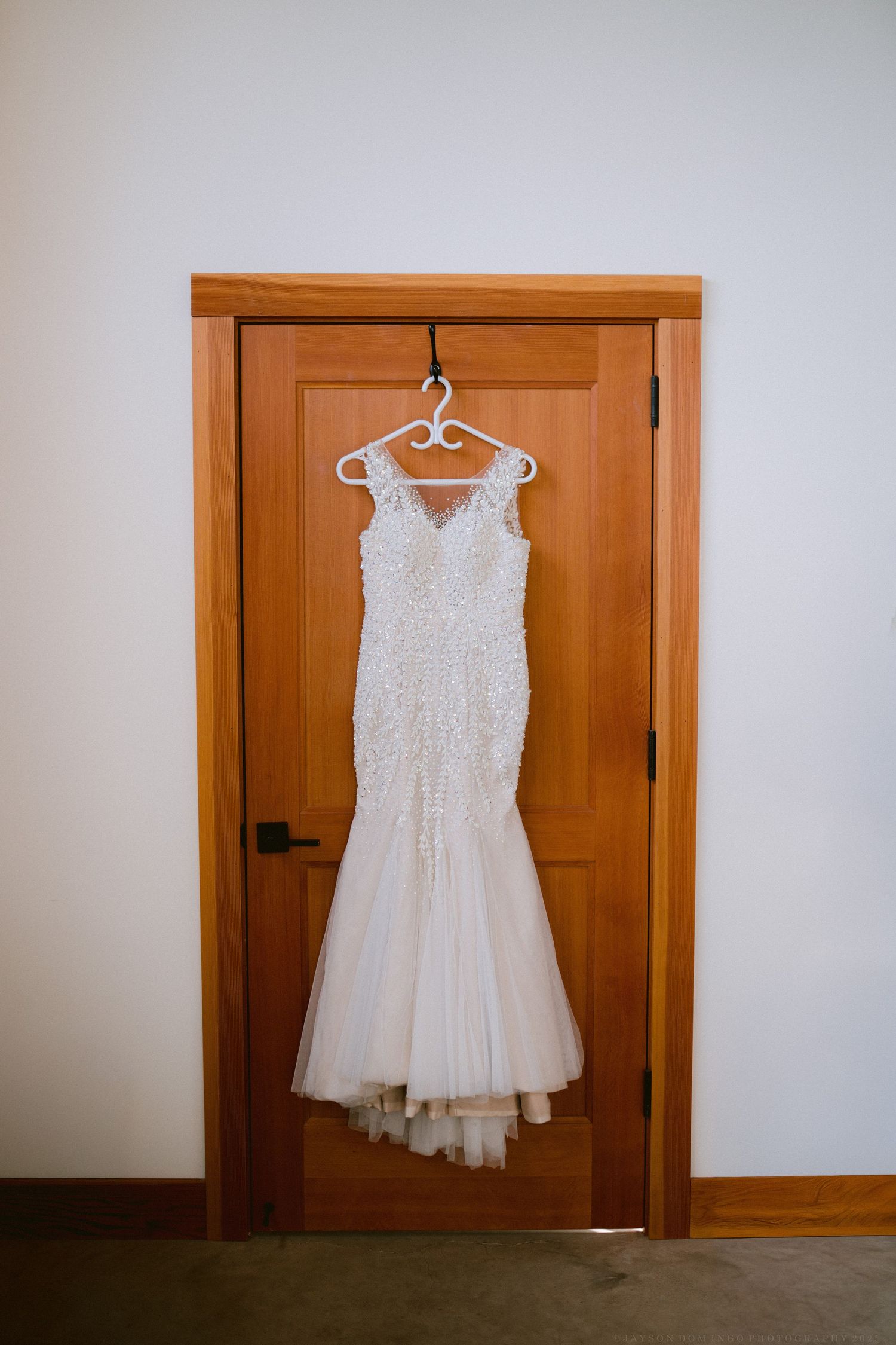 A white lace wedding dress hangs on a wooden hanger against a wooden door.