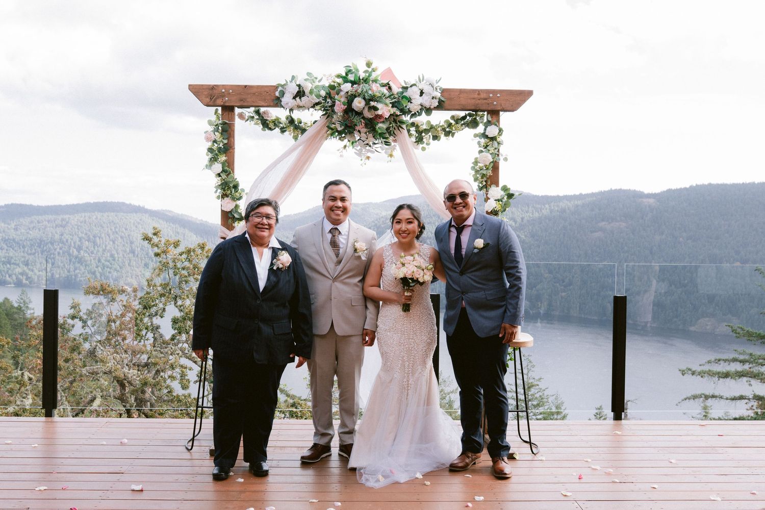 Wedding party gathered under floral arch on deck with mountain lake backdrop.