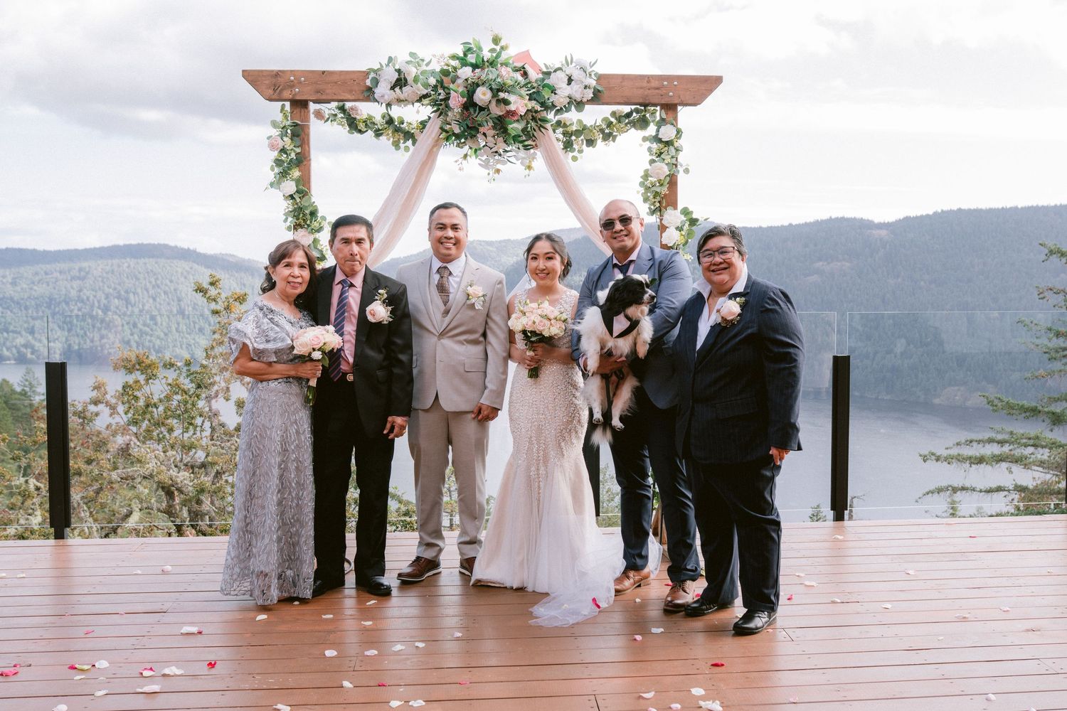 Wedding party poses together on wooden deck overlooking scenic mountain lake view with floral arch decor.