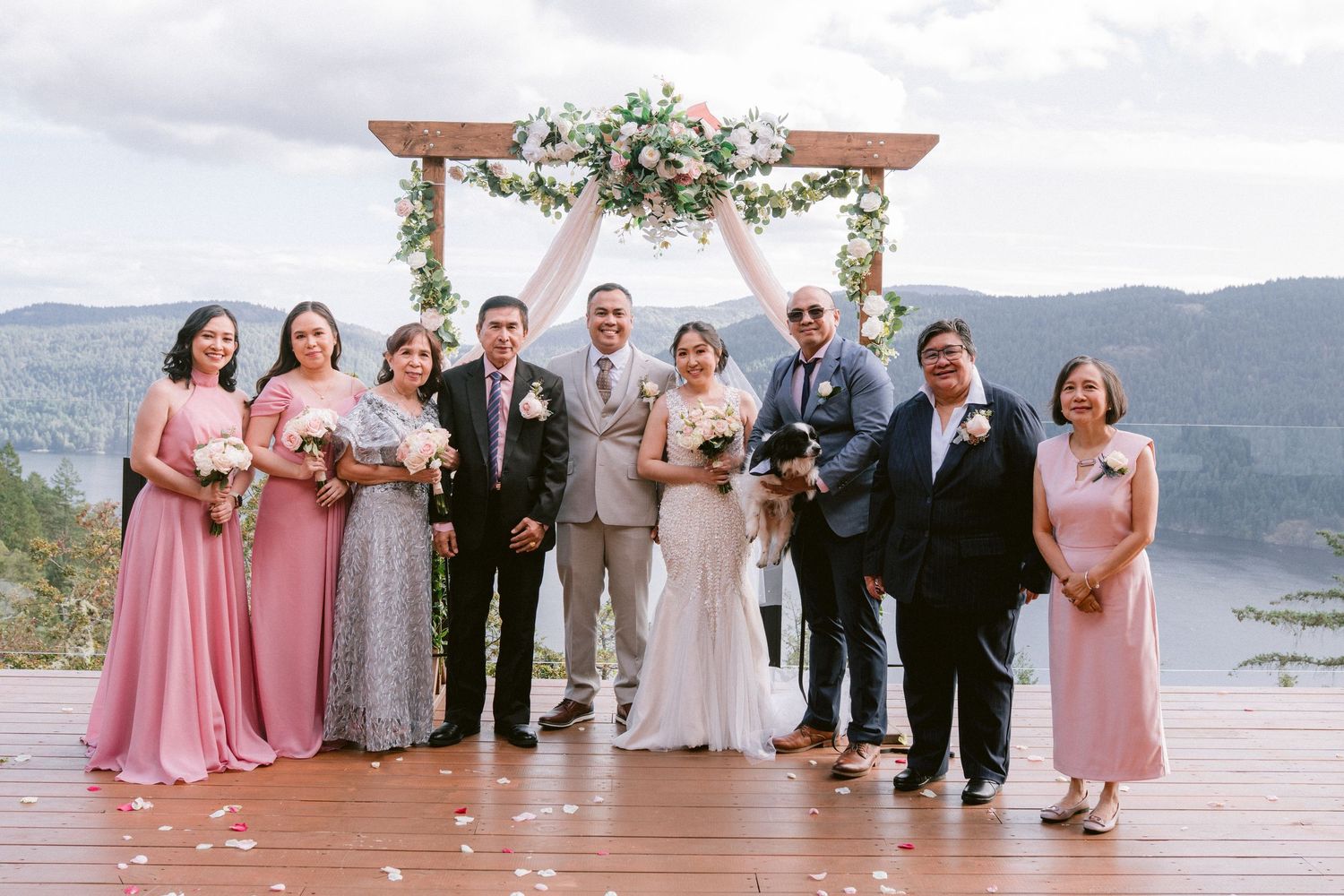 A wedding party stands on an outdoor deck with mountain views under a wooden arch decorated with white flowers.