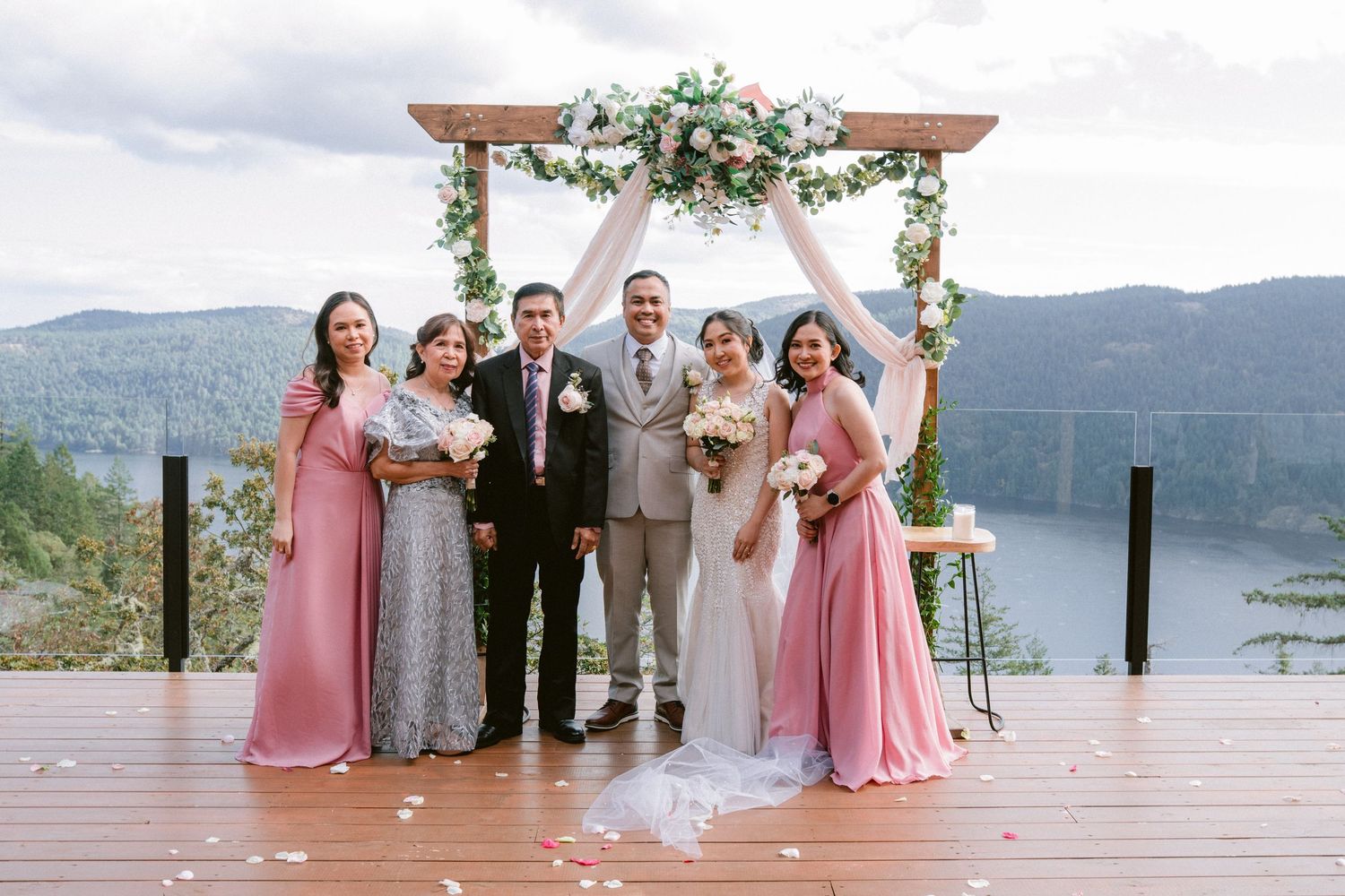 Wedding party poses on a lakeside dock with mountain views and floral arch decorations.