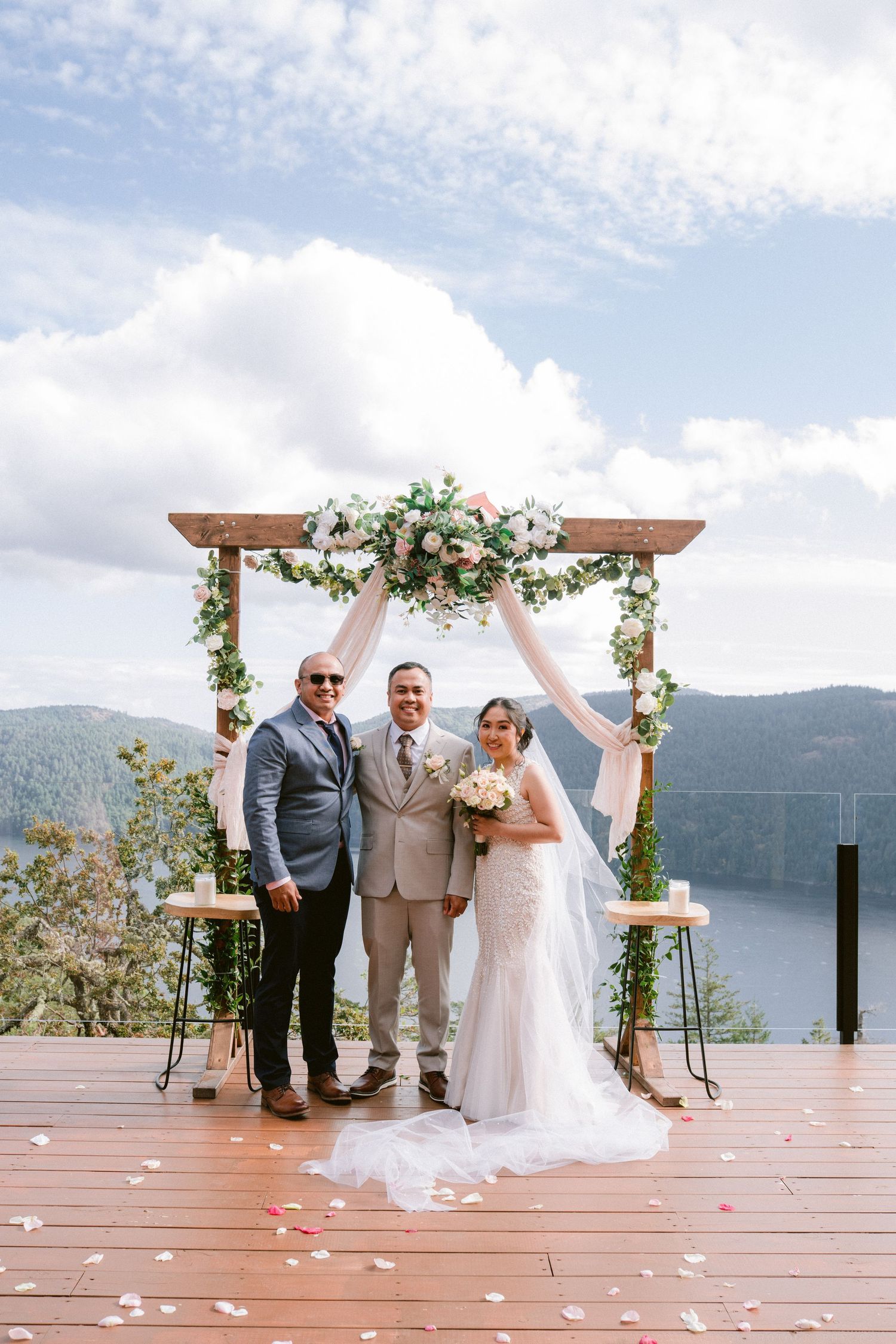 Outdoor lakeside wedding ceremony with wooden deck and floral arch overlooking mountain views.