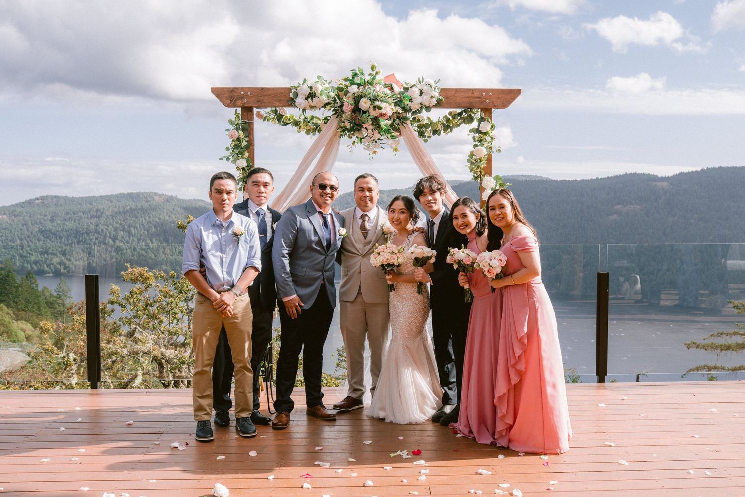 Wedding party poses on scenic outdoor deck overlooking mountain lake and forest landscape on sunny day.