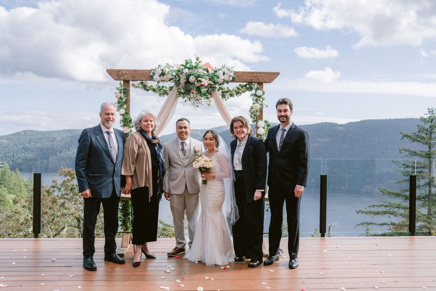 Wedding party poses under floral arch on mountain deck with scenic lake and mountain backdrop.