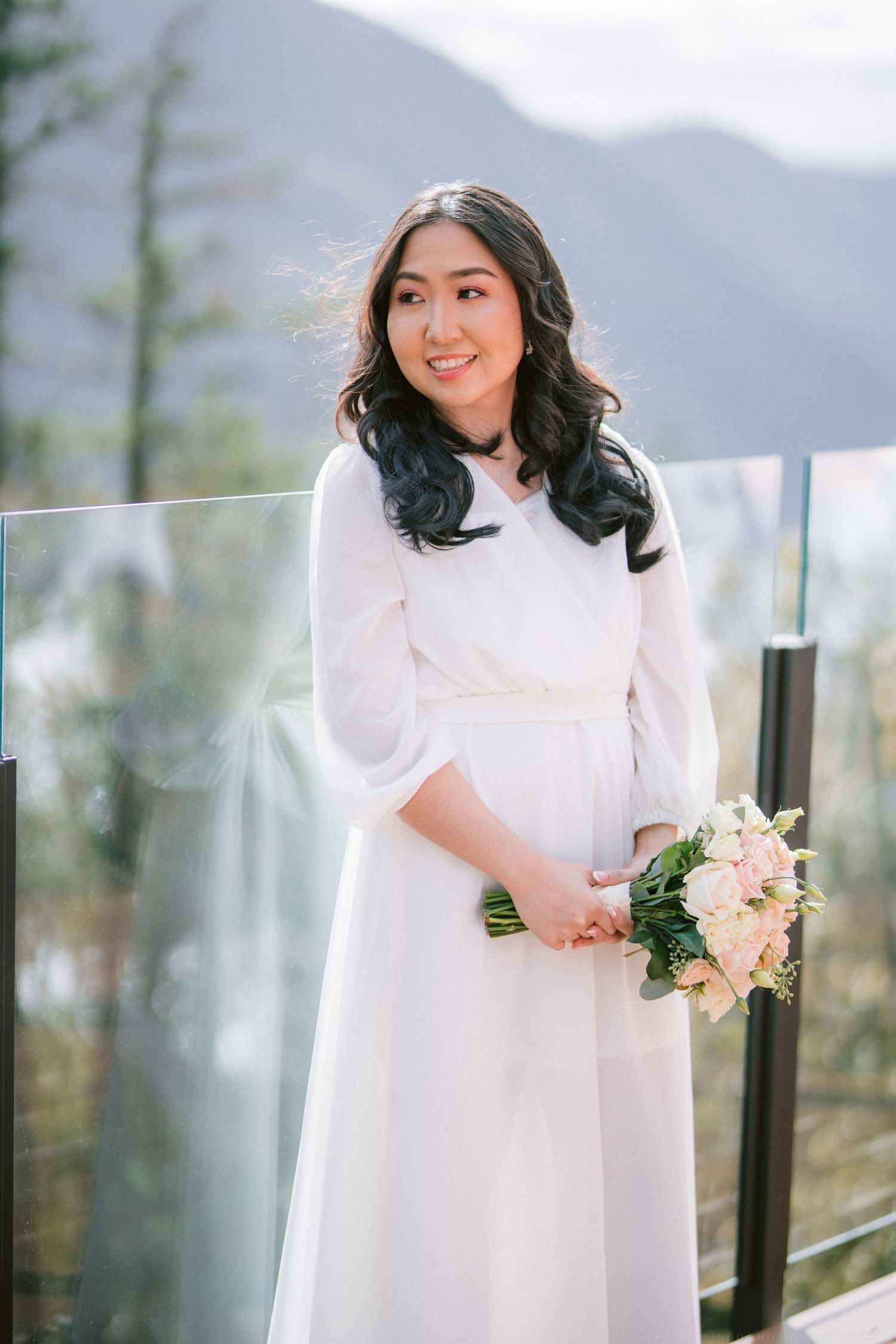 A bride in a flowing white dress holds a pastel bouquet while posing against a mountain backdrop.