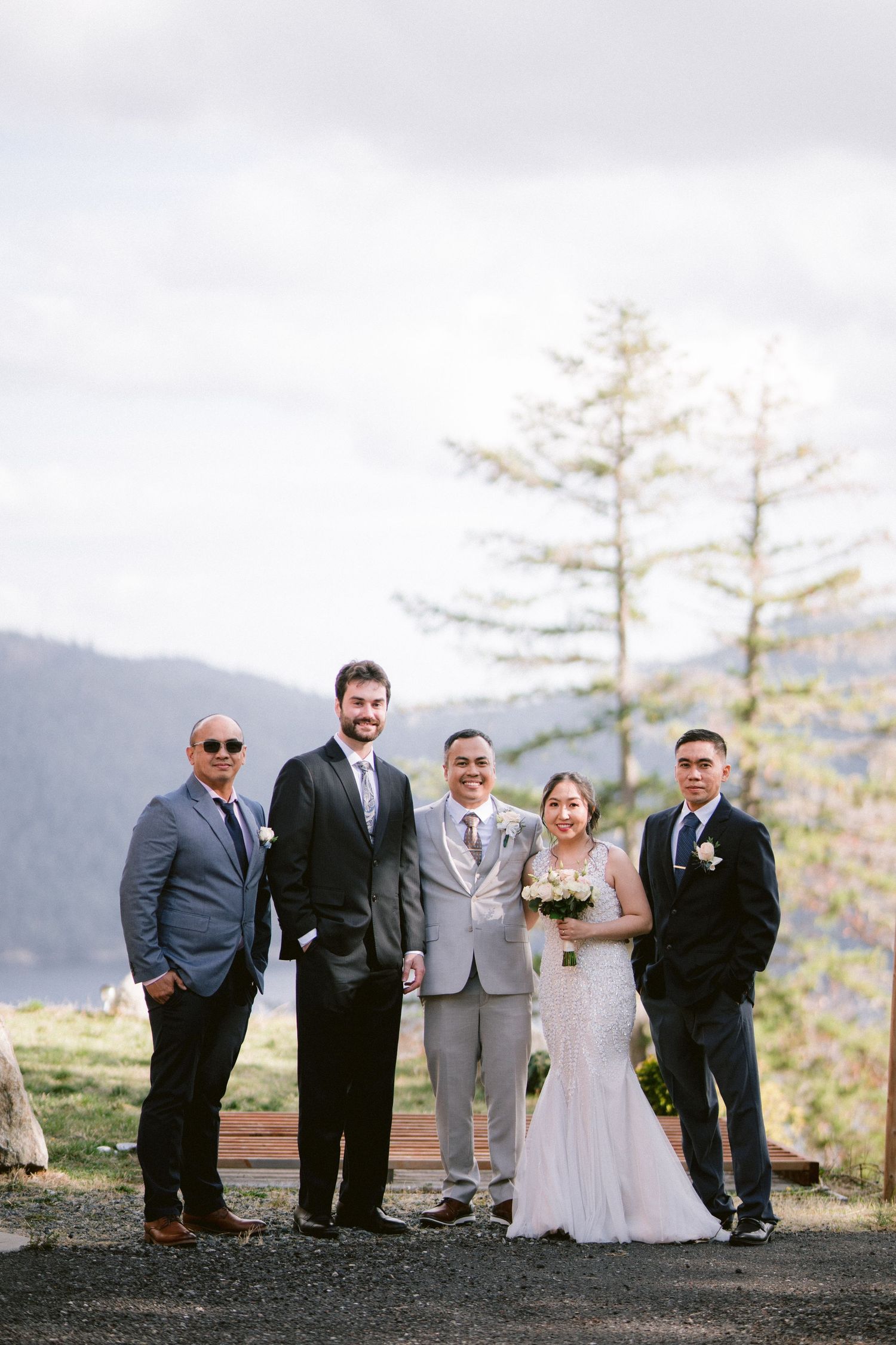 A wedding party stands together against a scenic mountain backdrop with tall evergreen trees in the autumn light.