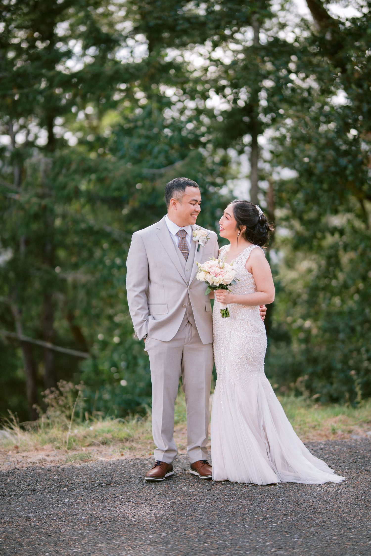 A wedding couple shares a romantic moment on a gravel path surrounded by trees.