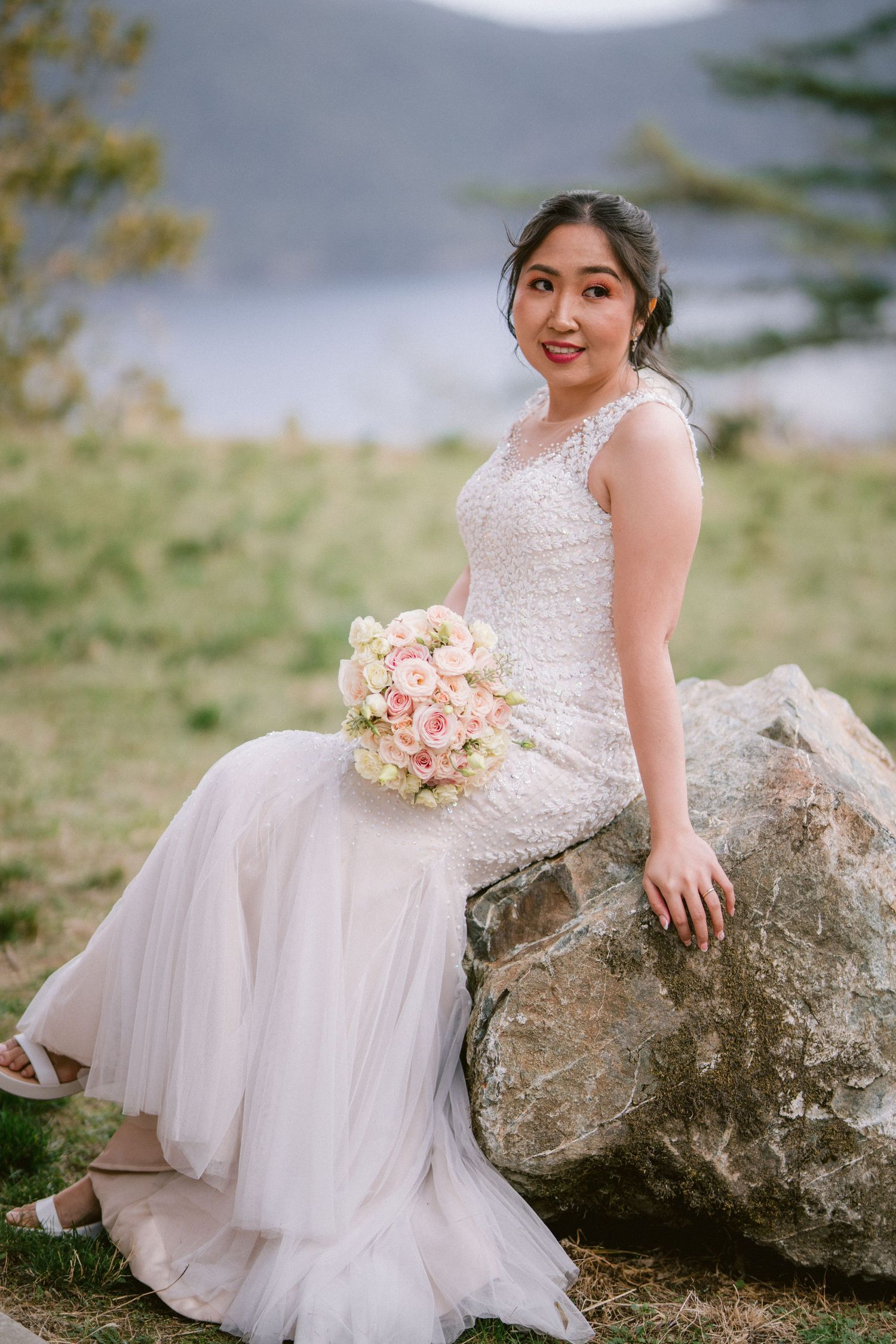 A bride in a white lace dress sits on large rocks with a pink bouquet.