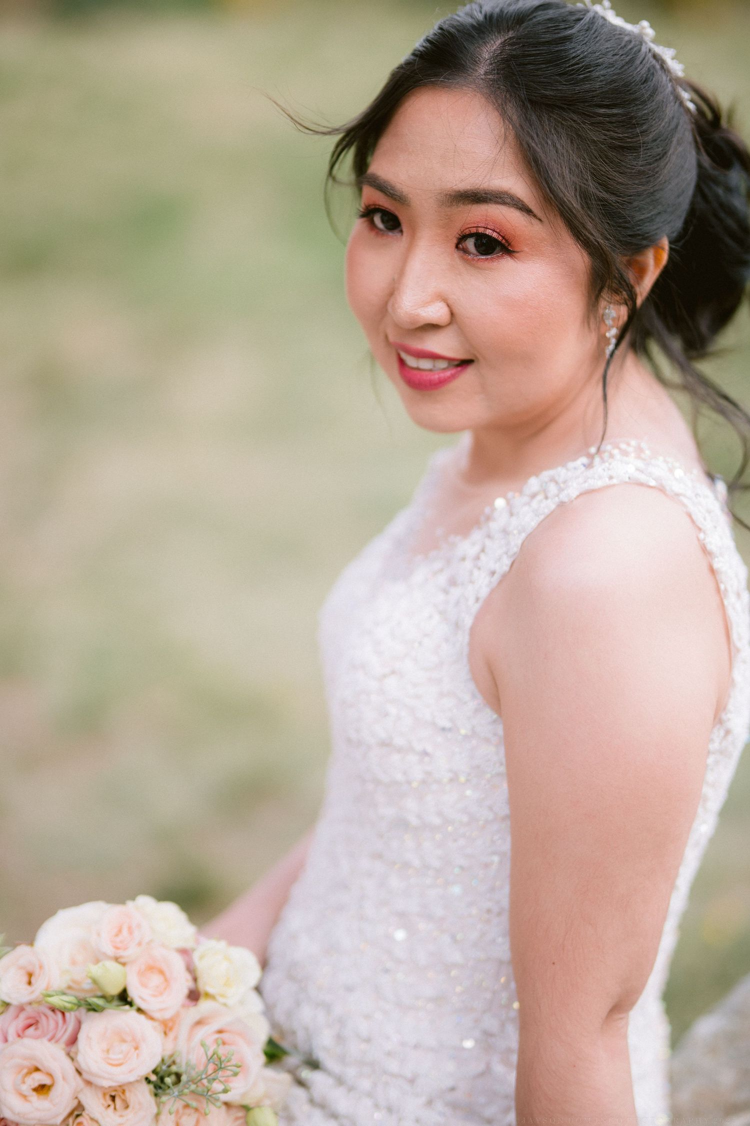 A bride in a white lace dress poses outdoors holding a soft pink and peach rose bouquet.