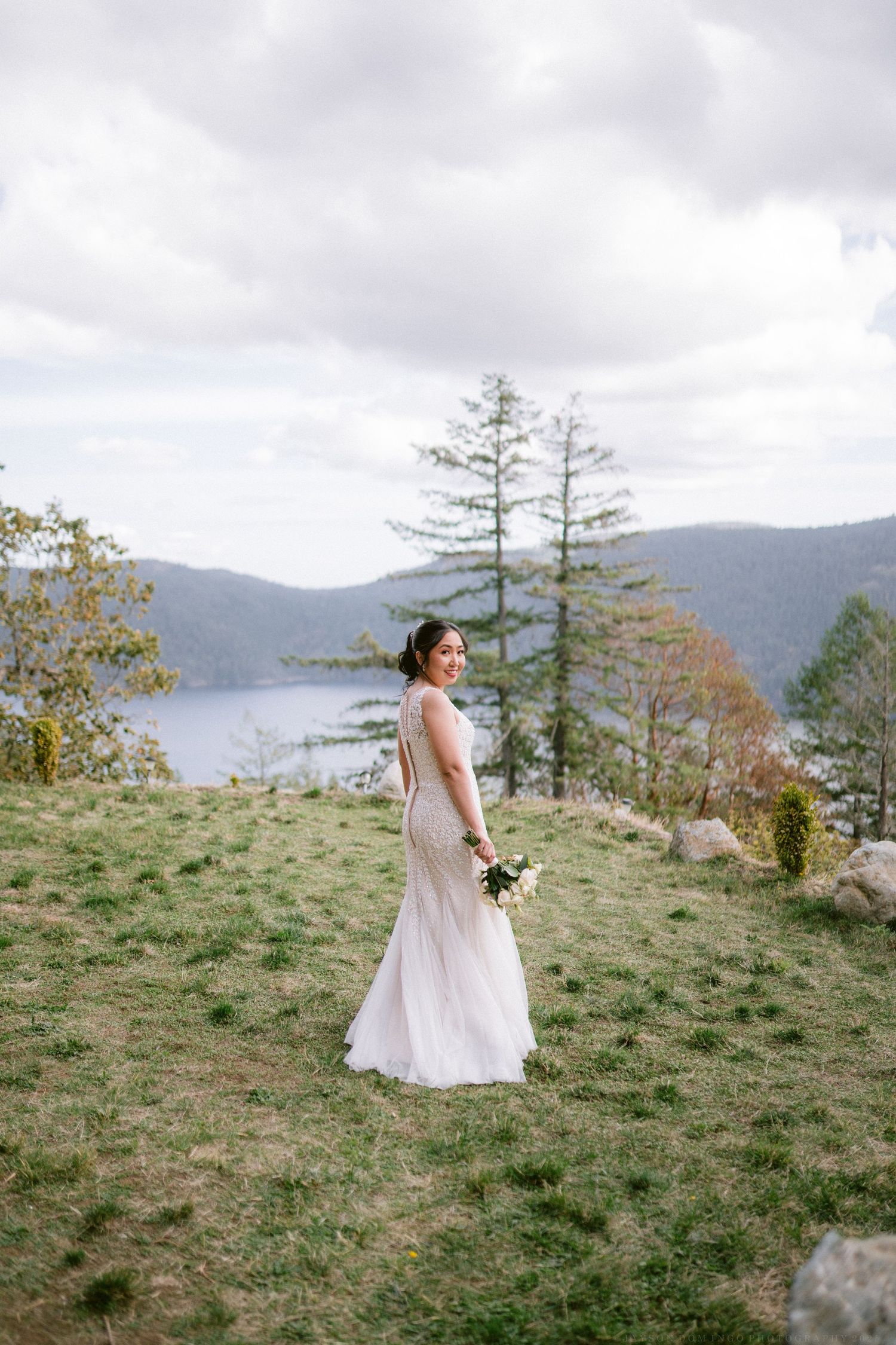 A bride in a fitted lace gown stands on a hillside overlooking a serene mountain lake.