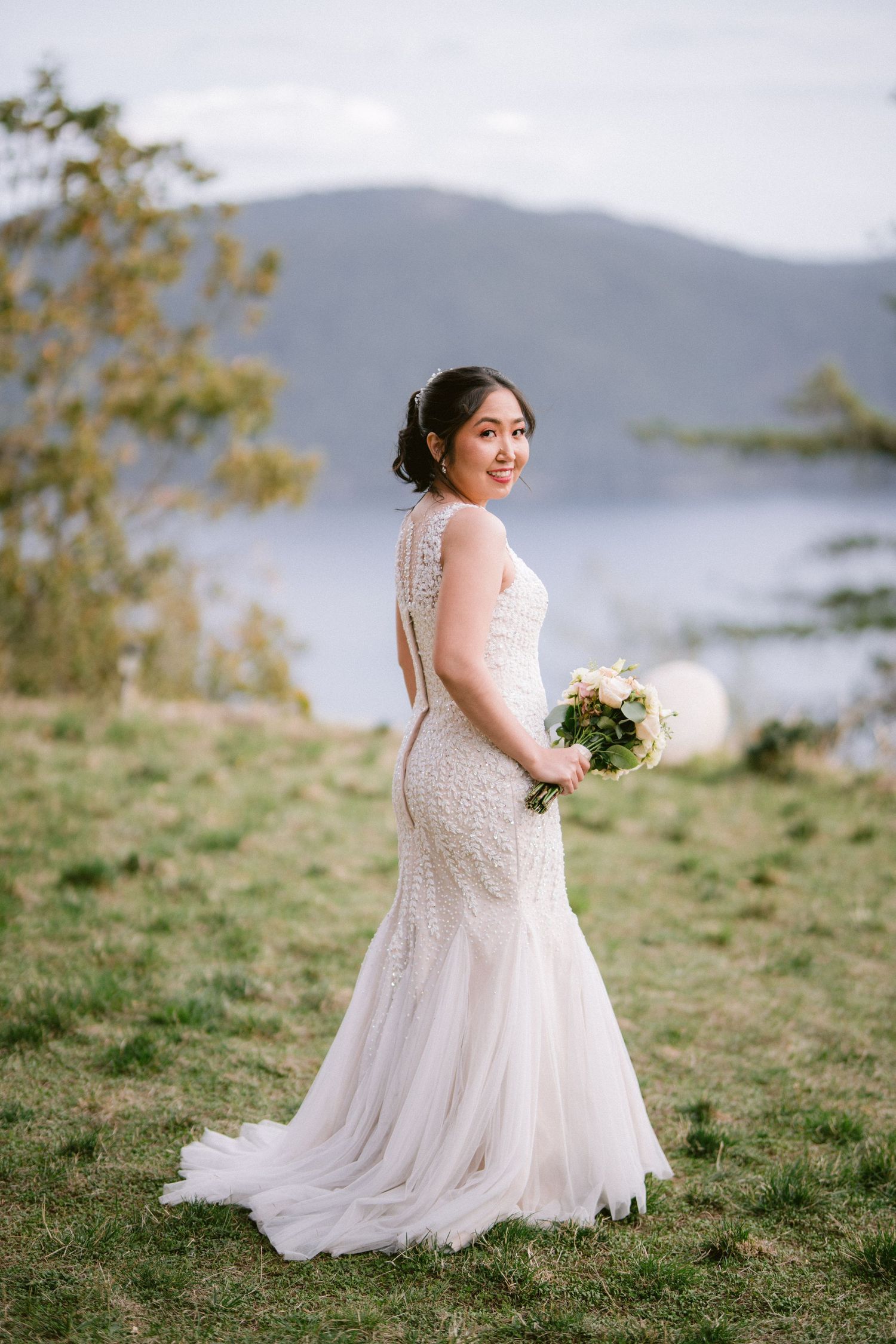 A bride in a flowing white dress poses on a grassy hillside with mountains in the background.