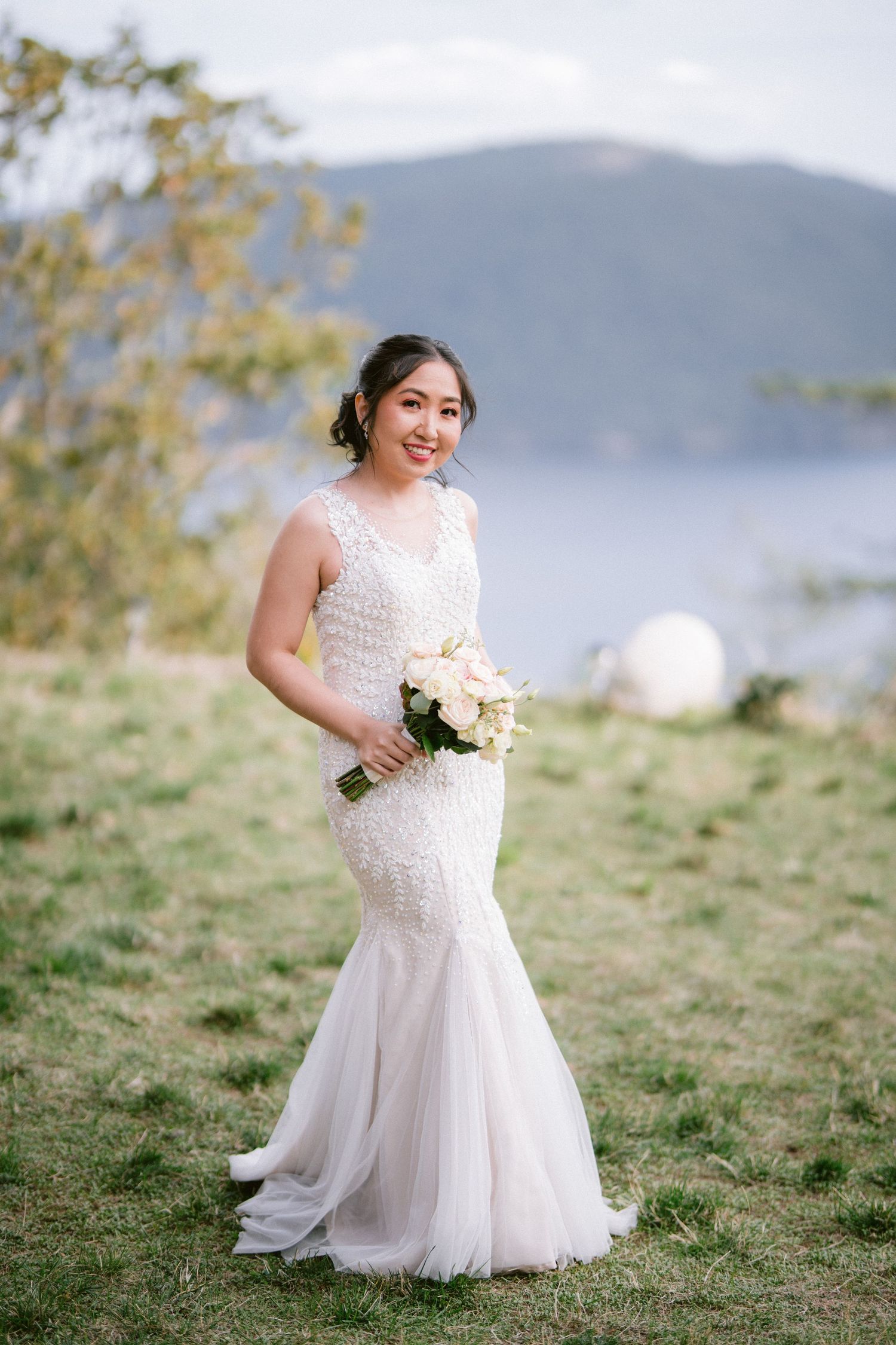 A bride in a white lace wedding dress poses on a grassy hillside with mountains in the background.