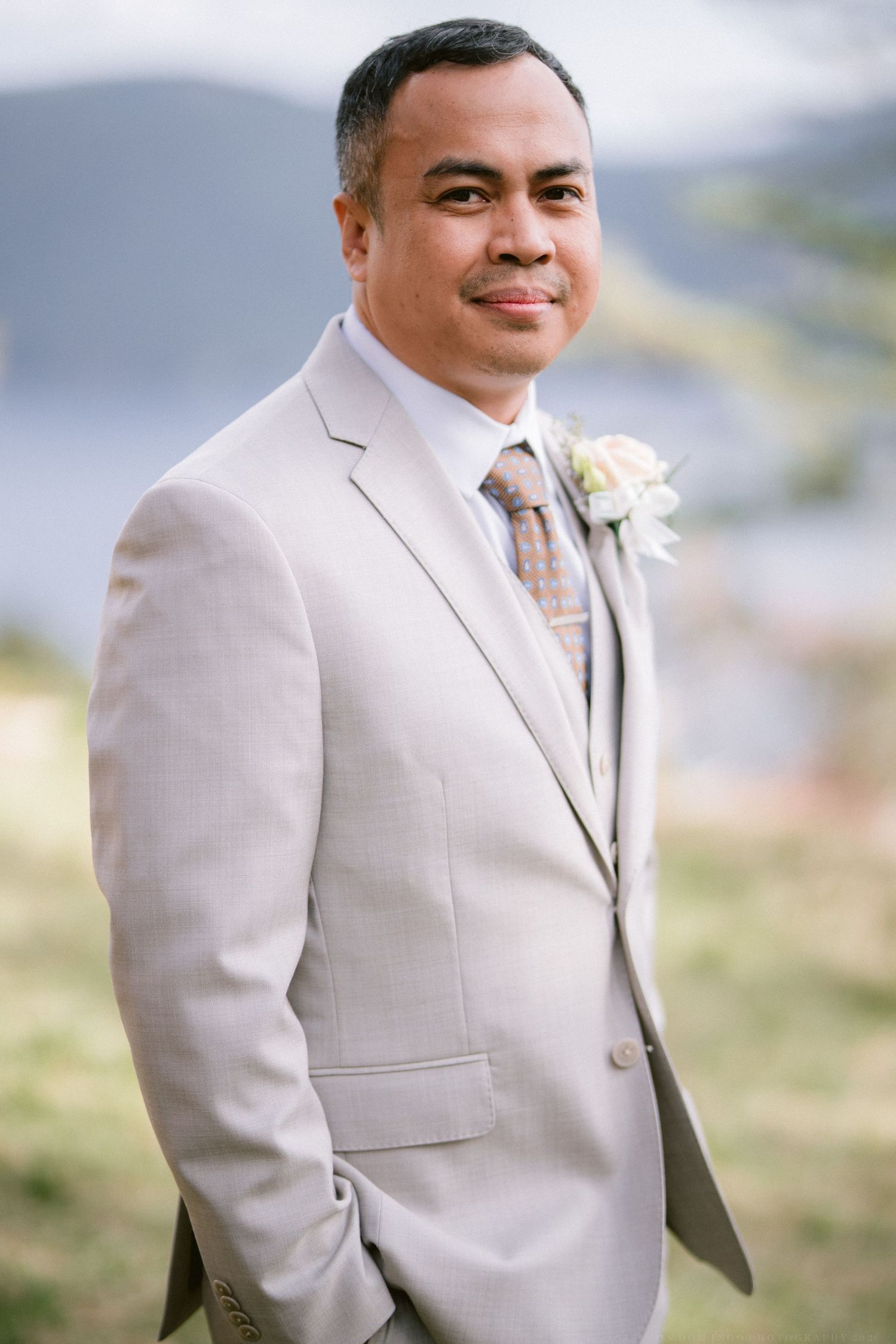 A groom in a light beige suit smiles warmly while standing outdoors on his wedding day.
