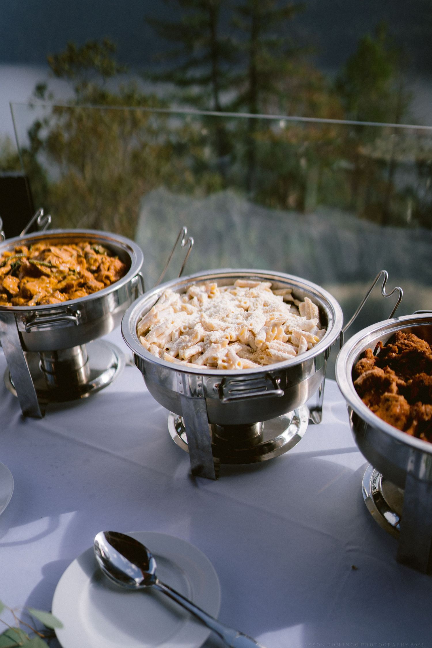Three chafing dishes filled with catered food sit on a white tablecloth at an outdoor reception.