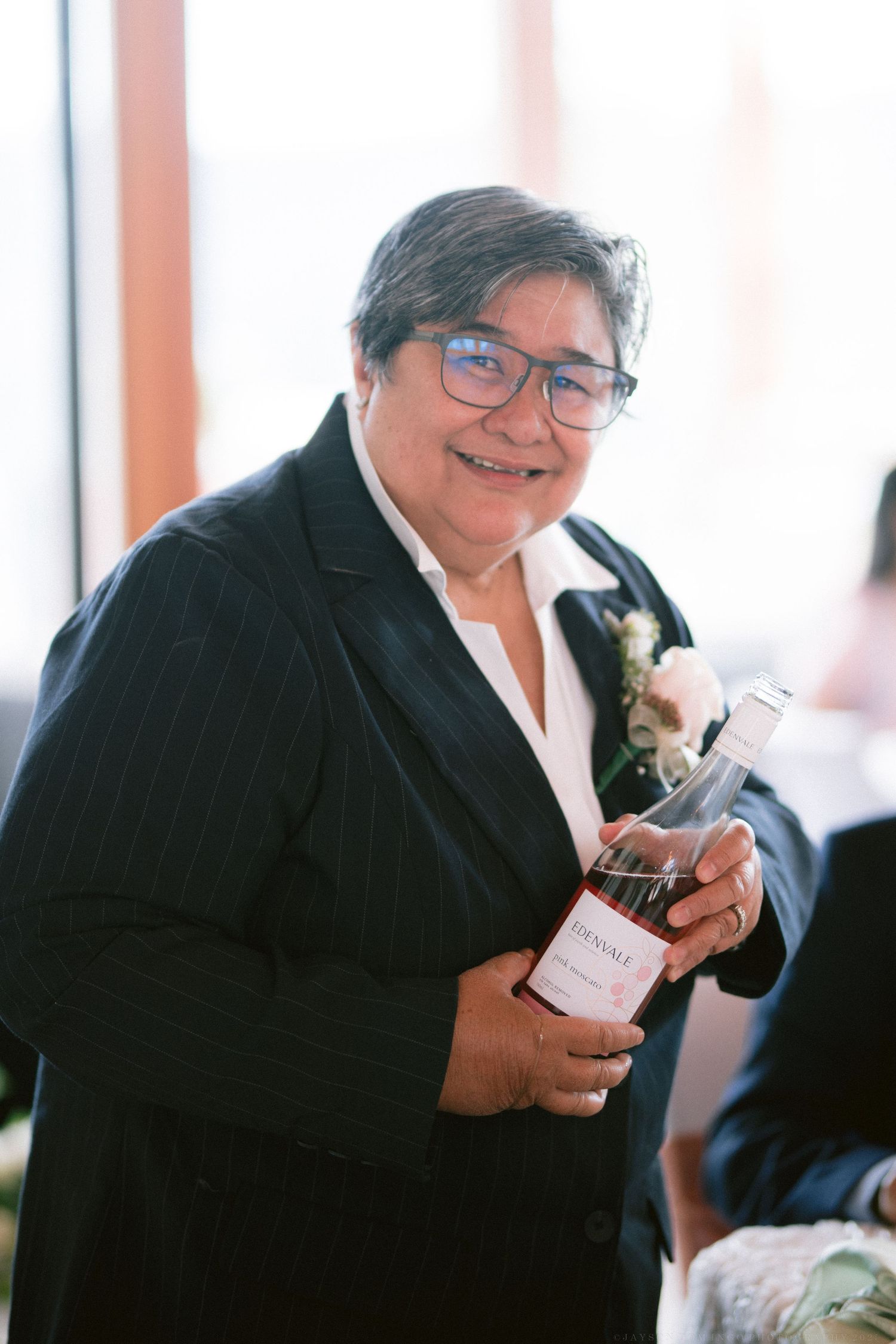 A guest in formal attire holds a wine bottle during the wedding celebration.