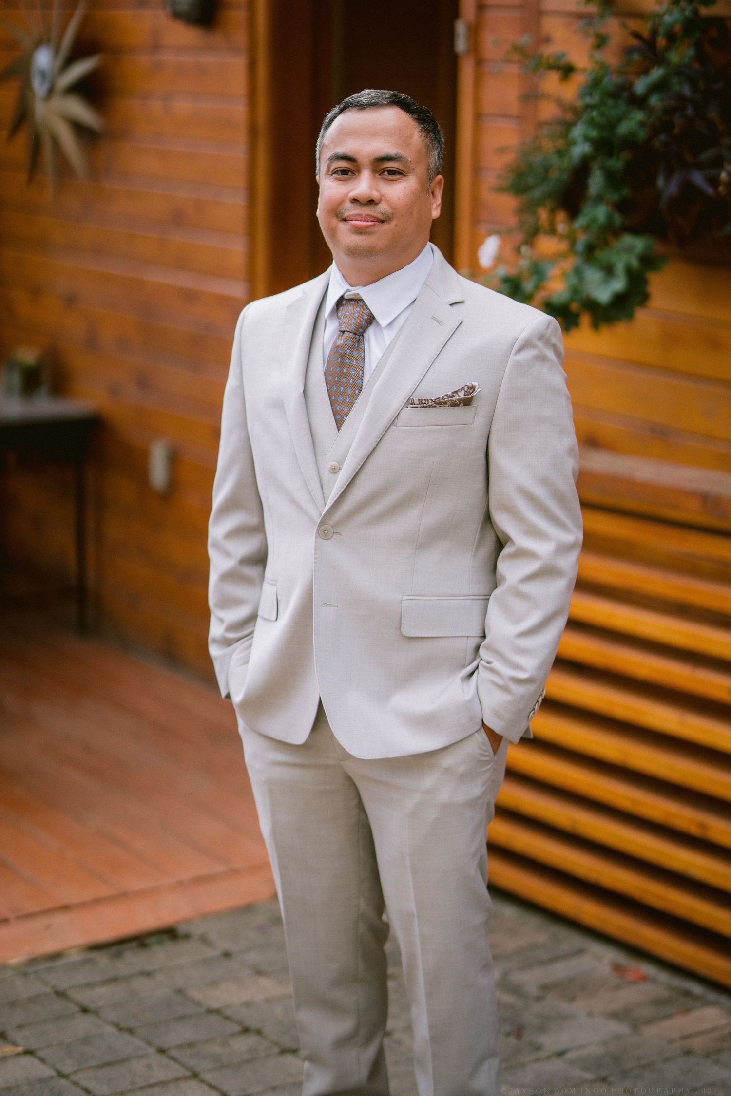 A person in a light beige suit and tie stands confidently against a wooden wall exterior.