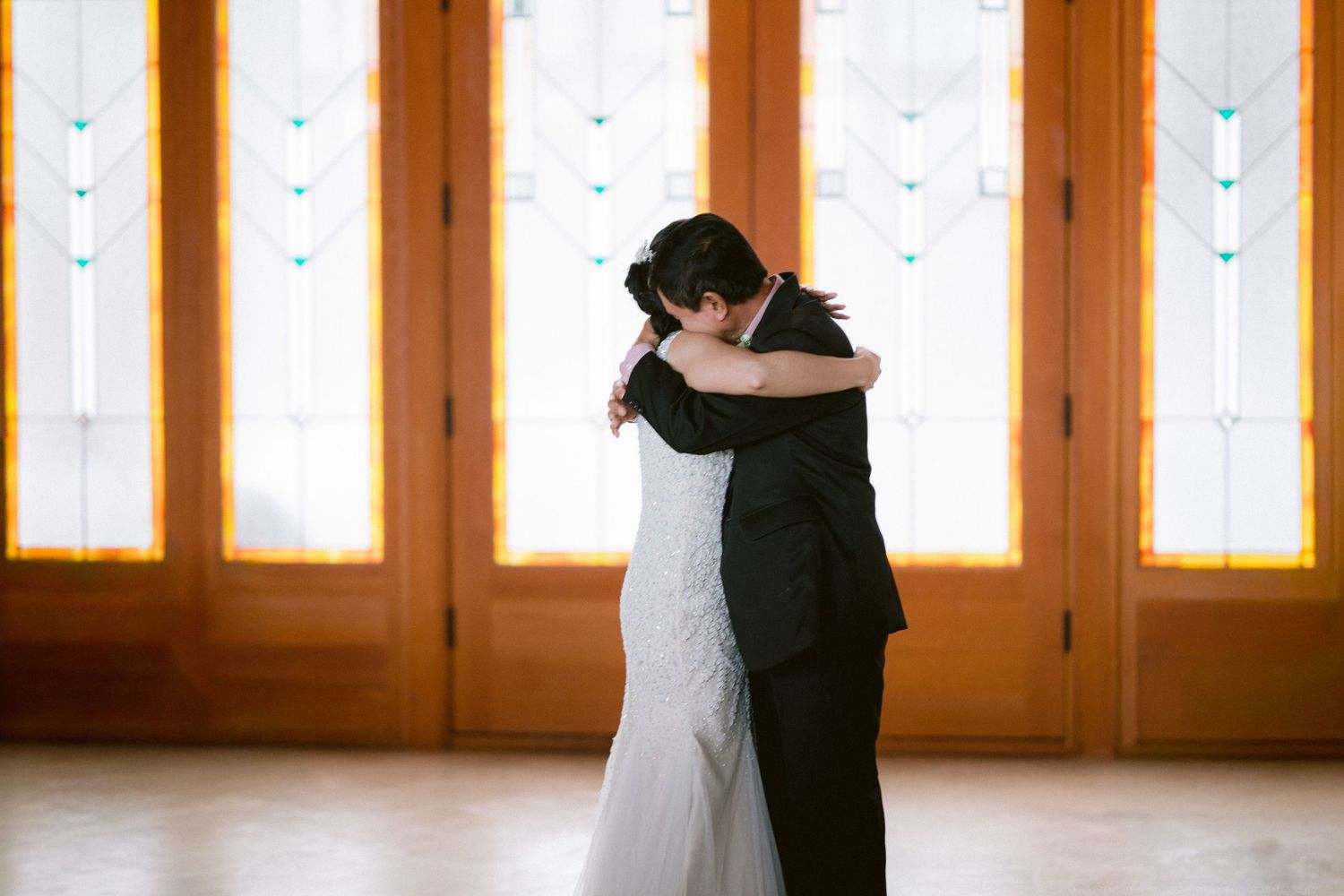 A couple in formal attire dances gracefully in a sunlit ballroom with wooden floors and large windows.
