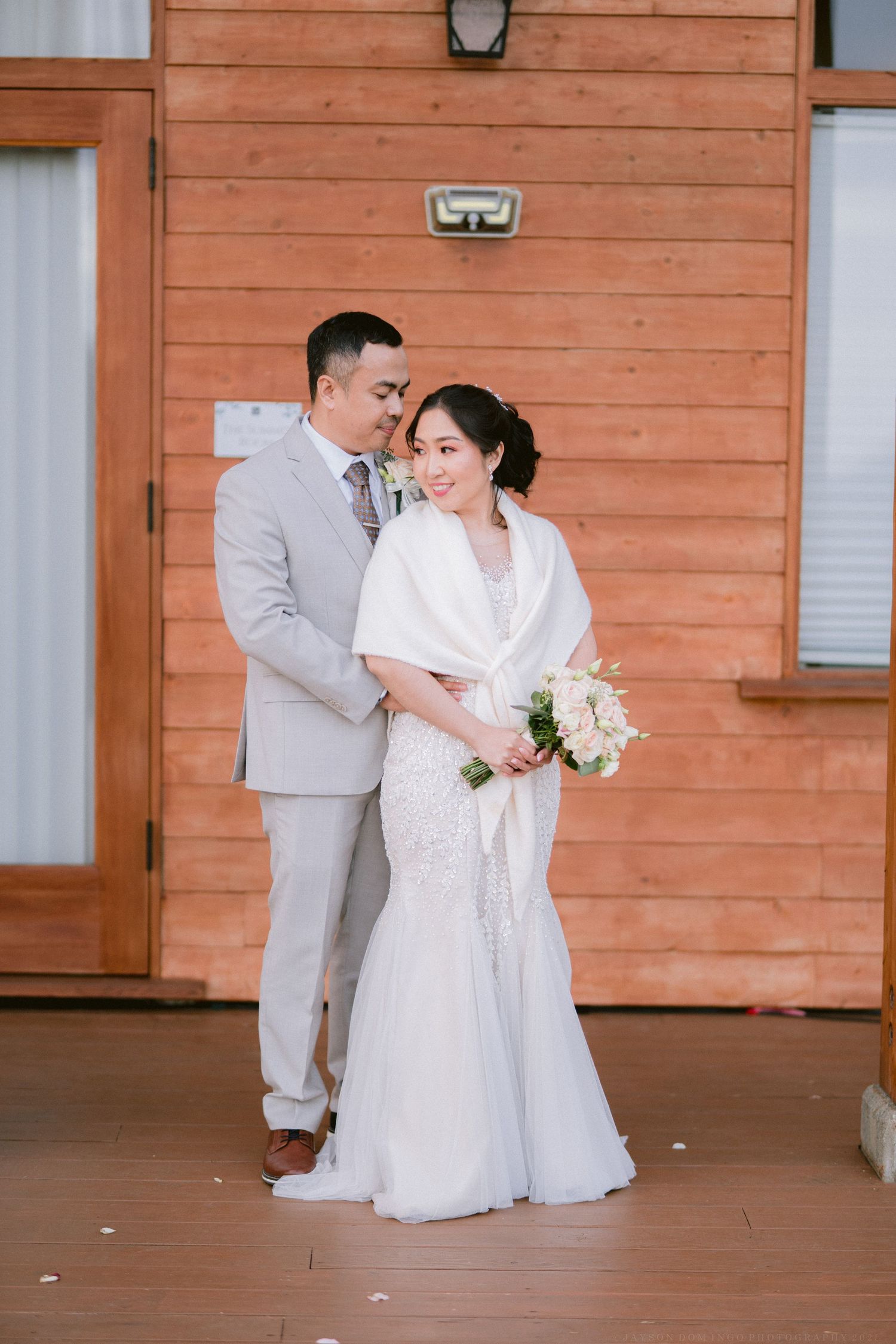 Couple poses against rustic orange wooden wall in elegant wedding attire.