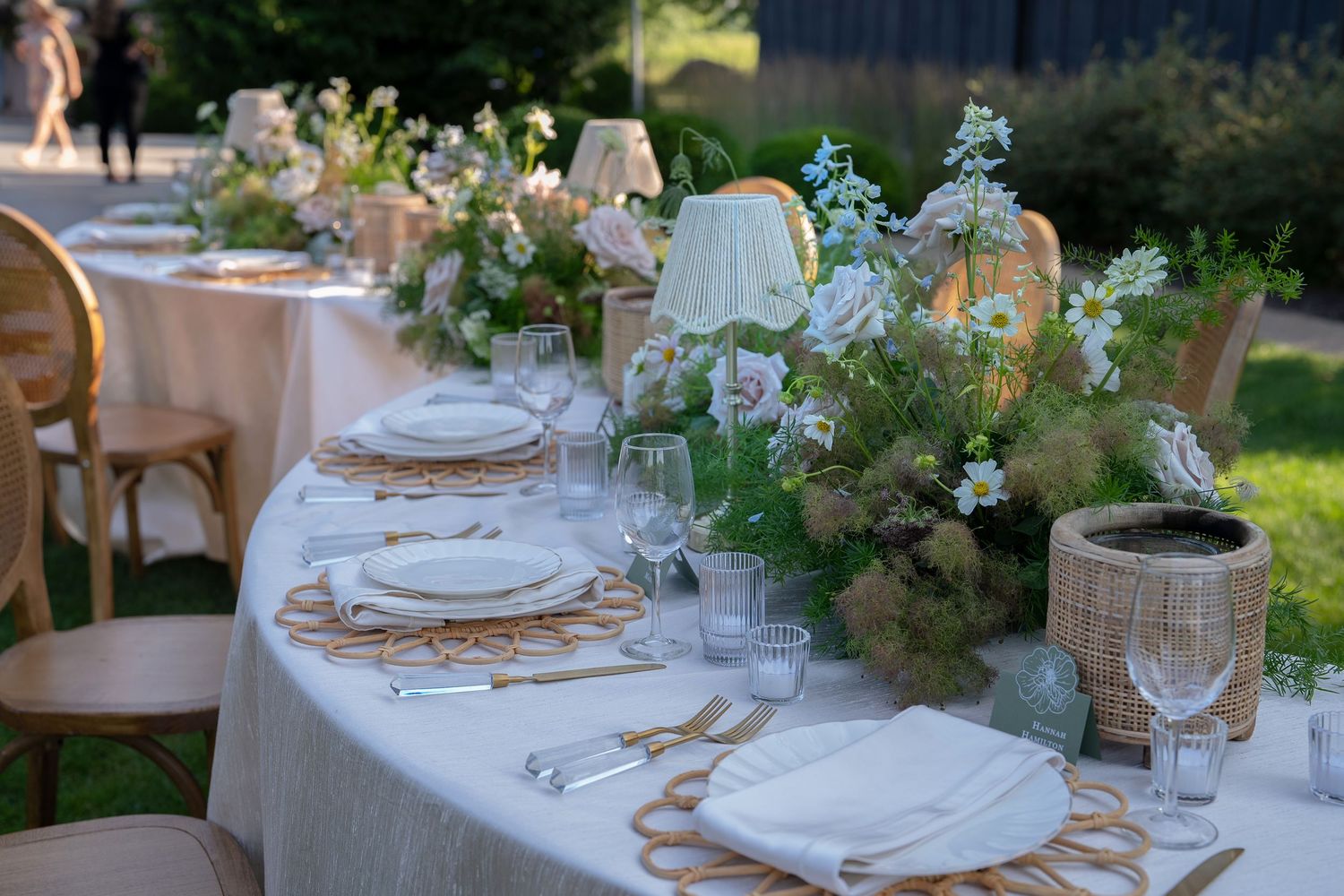 Elegant outdoor wedding reception tables decorated with white linens, floral arrangements and glassware in a garden setting.